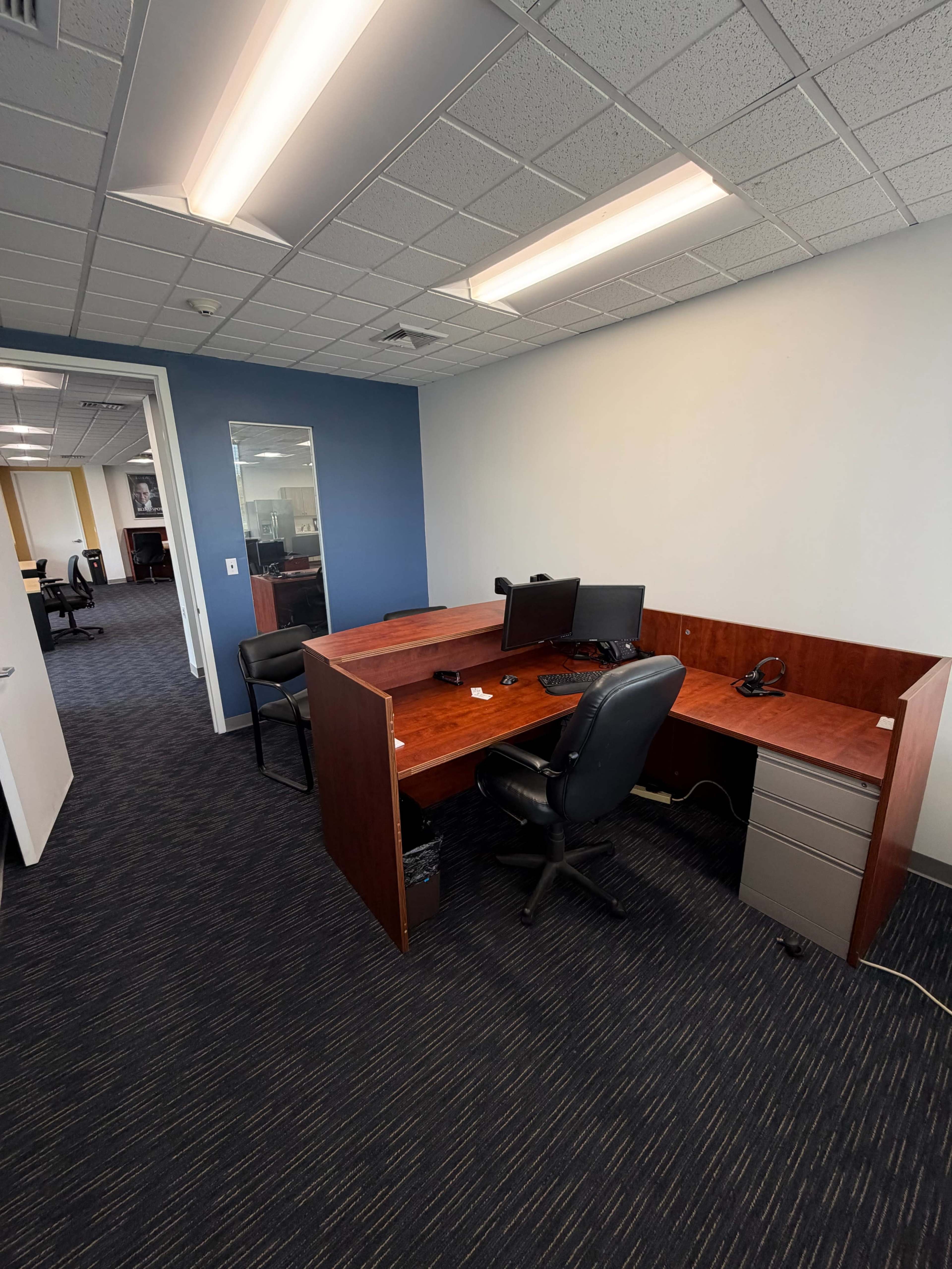 The image shows an empty office space with wooden desks, computer monitors, and a black office chair.