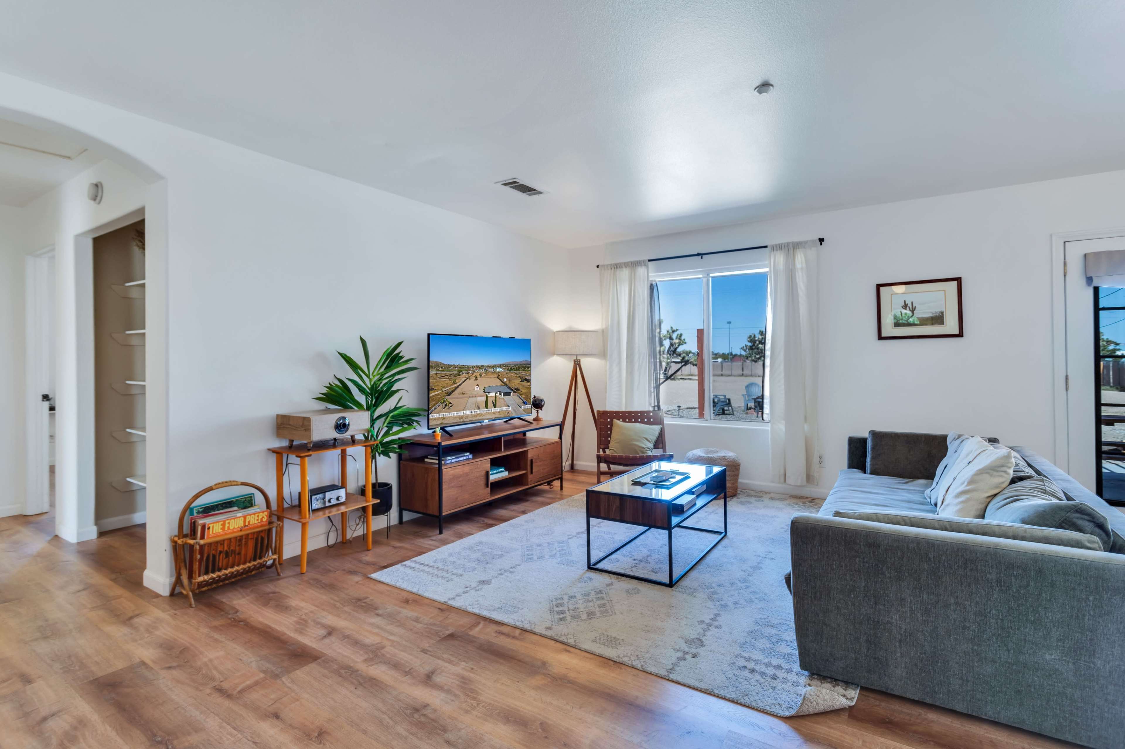 A modern living room features a large television, a comfortable sofa, a coffee table, and a window with sheer curtains allowing natural light.