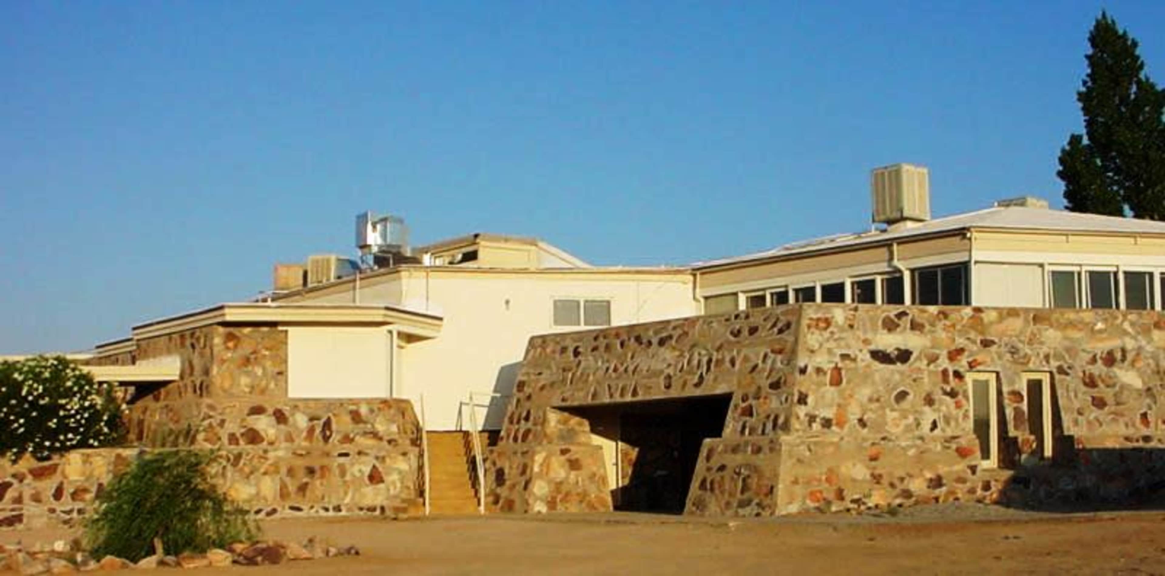 The image shows a modern building with a stone facade and flat roofs, surrounded by a dry landscape.