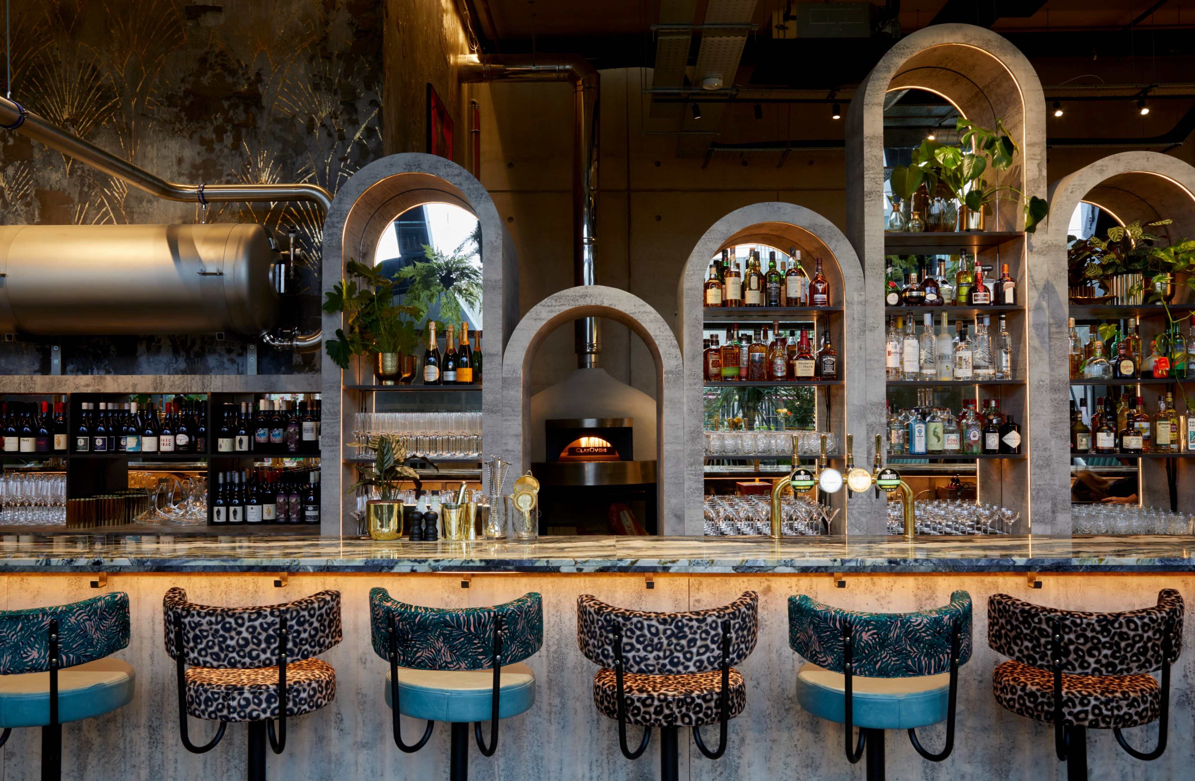 The image features a modern bar with a stone counter, a row of leopard-print barstools, and shelves lined with various bottles, complemented by greenery and an oven.