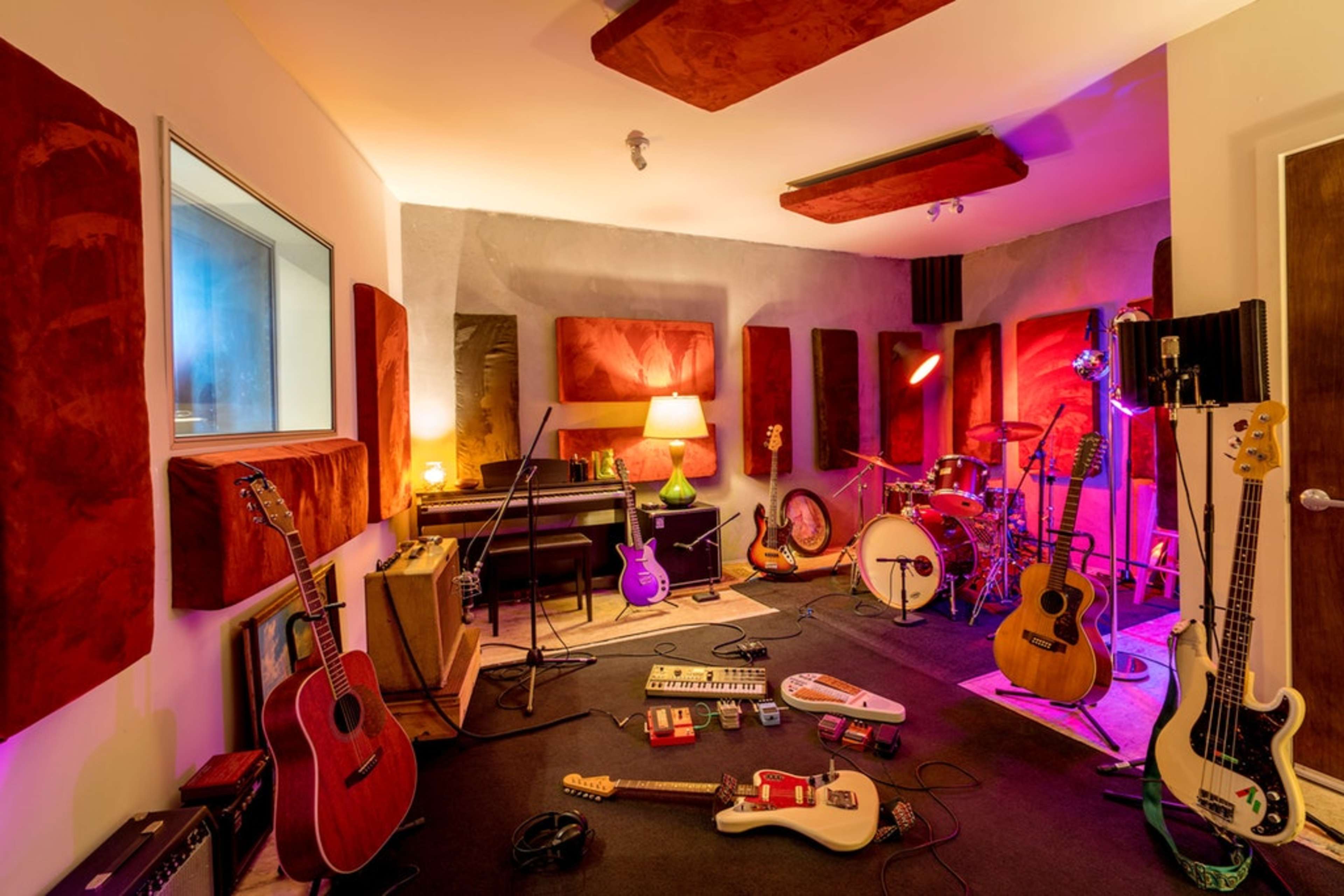 A music recording studio features various instruments scattered on the floor, soundproofing panels on the walls, and a drum set set up in the corner.