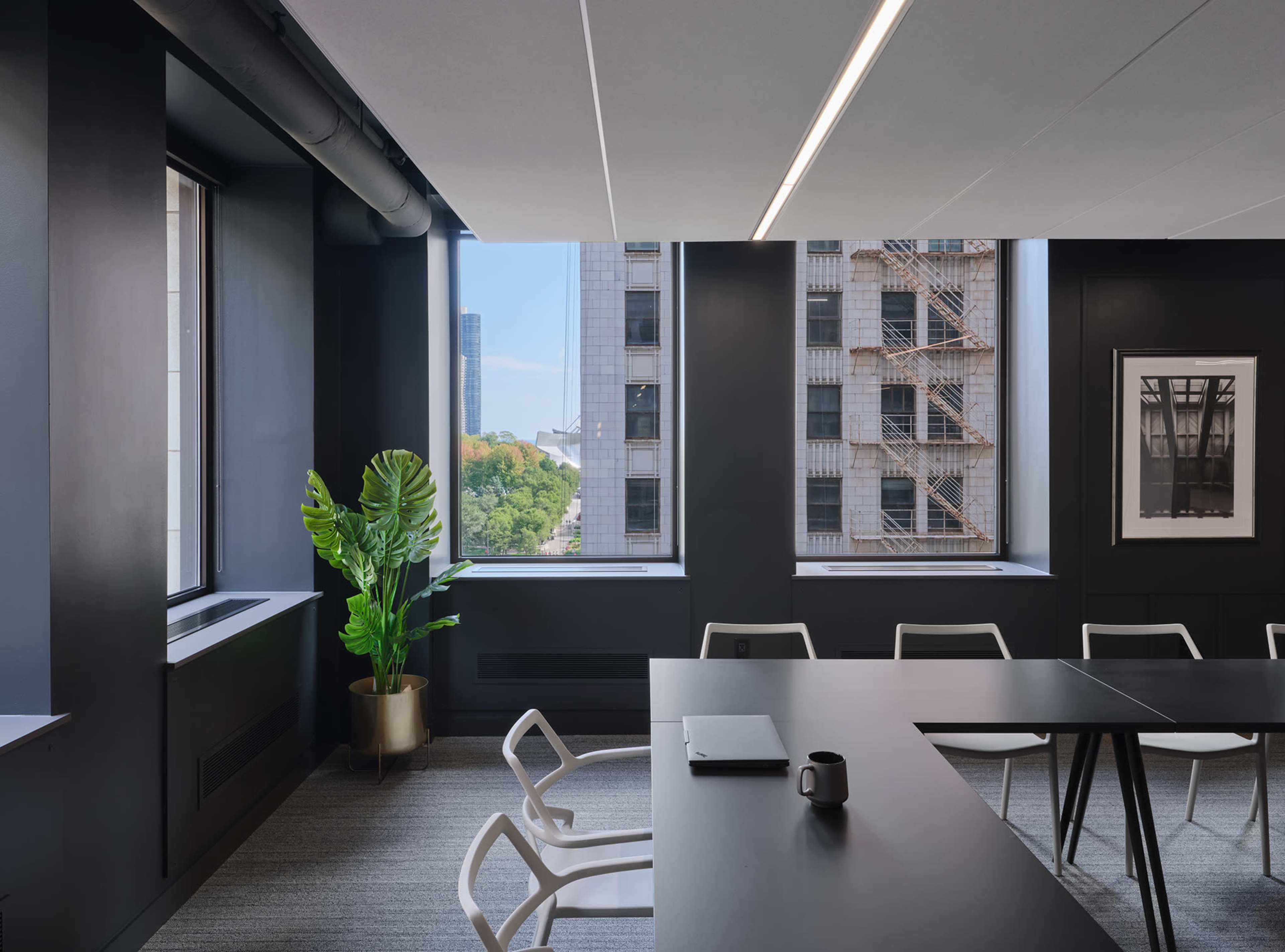 The image shows a modern office space with a large table, white chairs, and a potted plant near a window, revealing a view of city buildings outside.