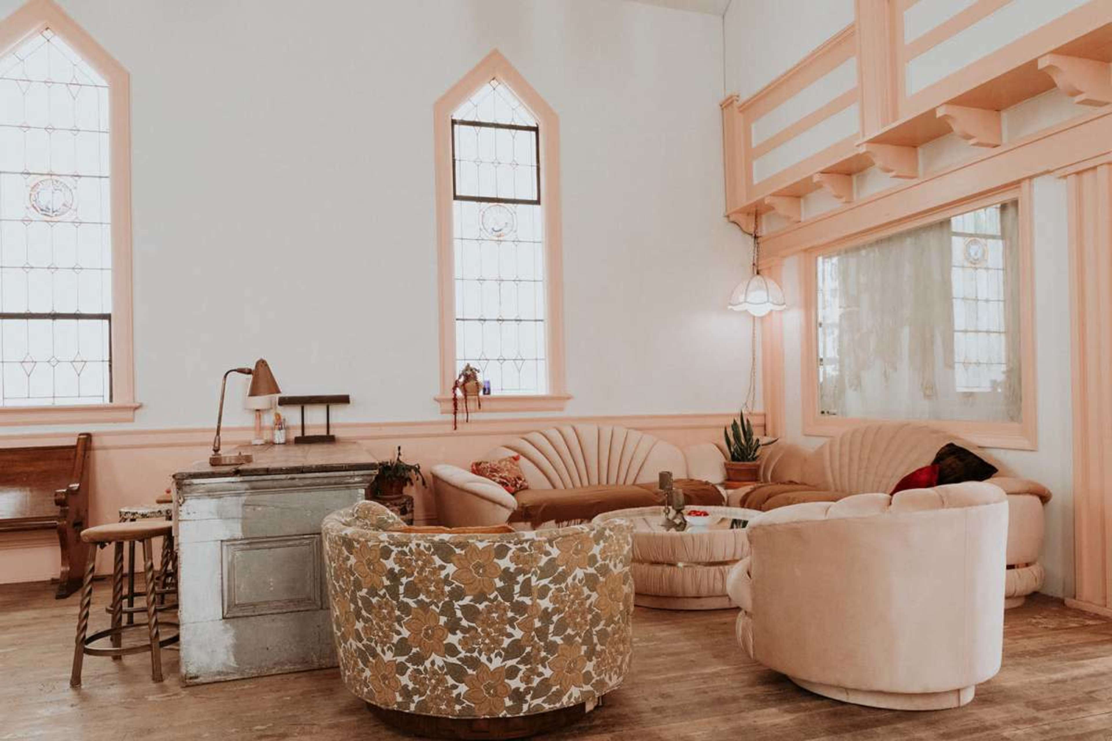 The image shows a bright room with vintage-style furniture, including a curved sofa and plush armchairs, against a backdrop of large stained glass windows.