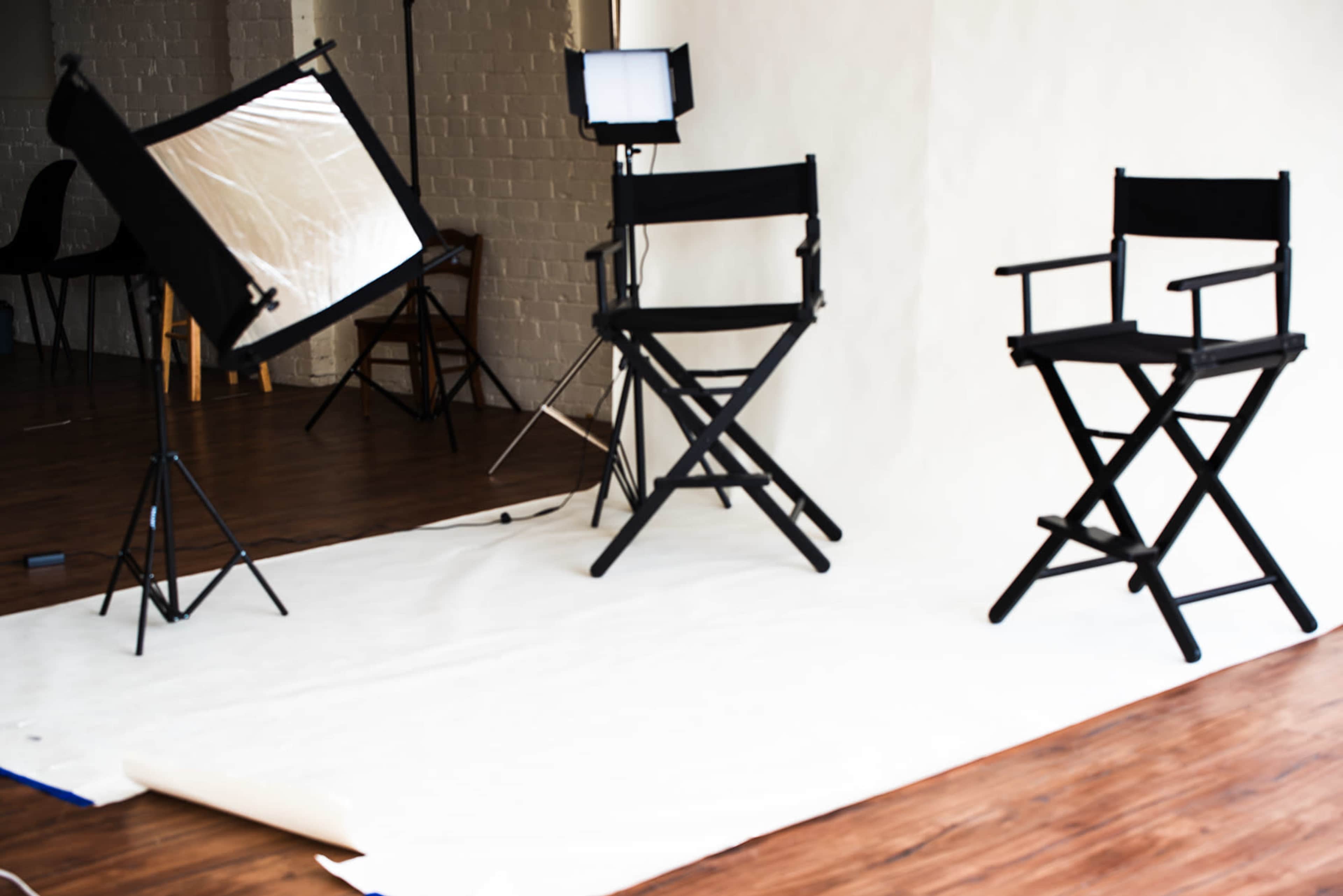 A photo studio setup features two black director's chairs, a white backdrop, and lighting equipment arranged on a wooden floor.