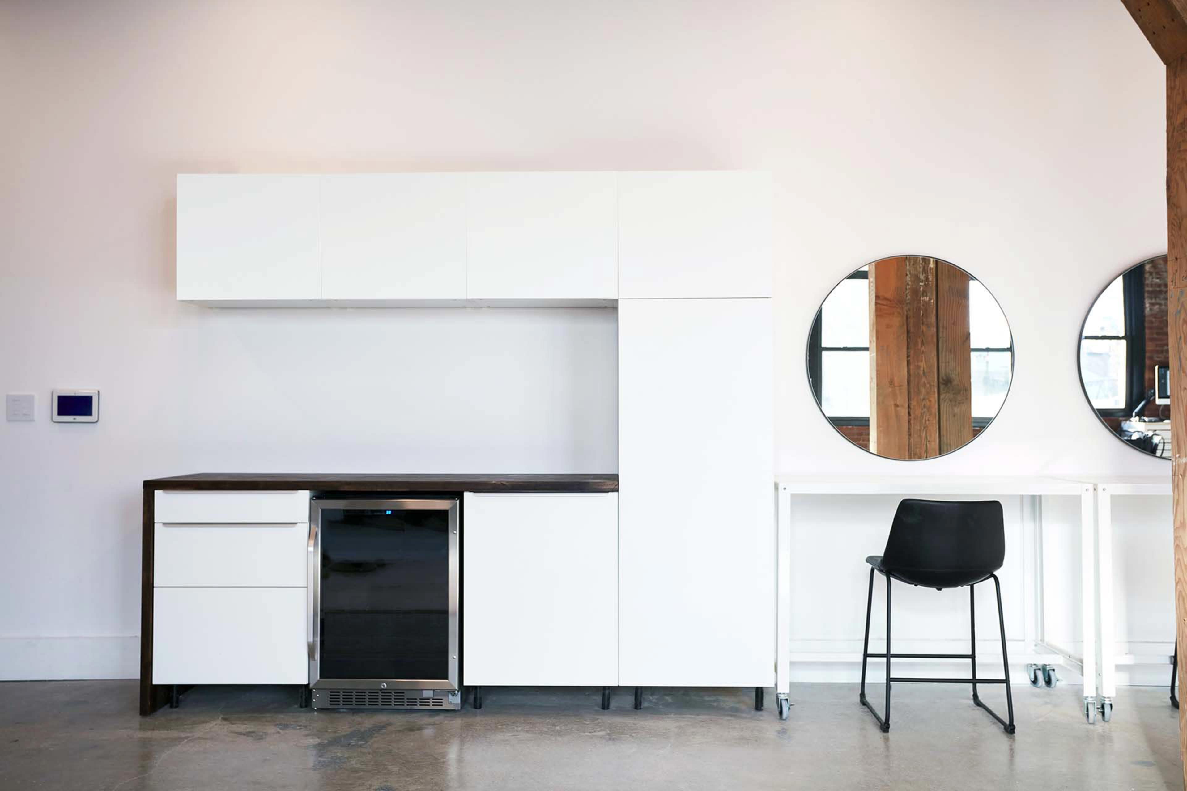 A modern kitchen setup features white cabinetry, a sleek refrigerator, and a round mirror above a minimalistic desk with a black chair.