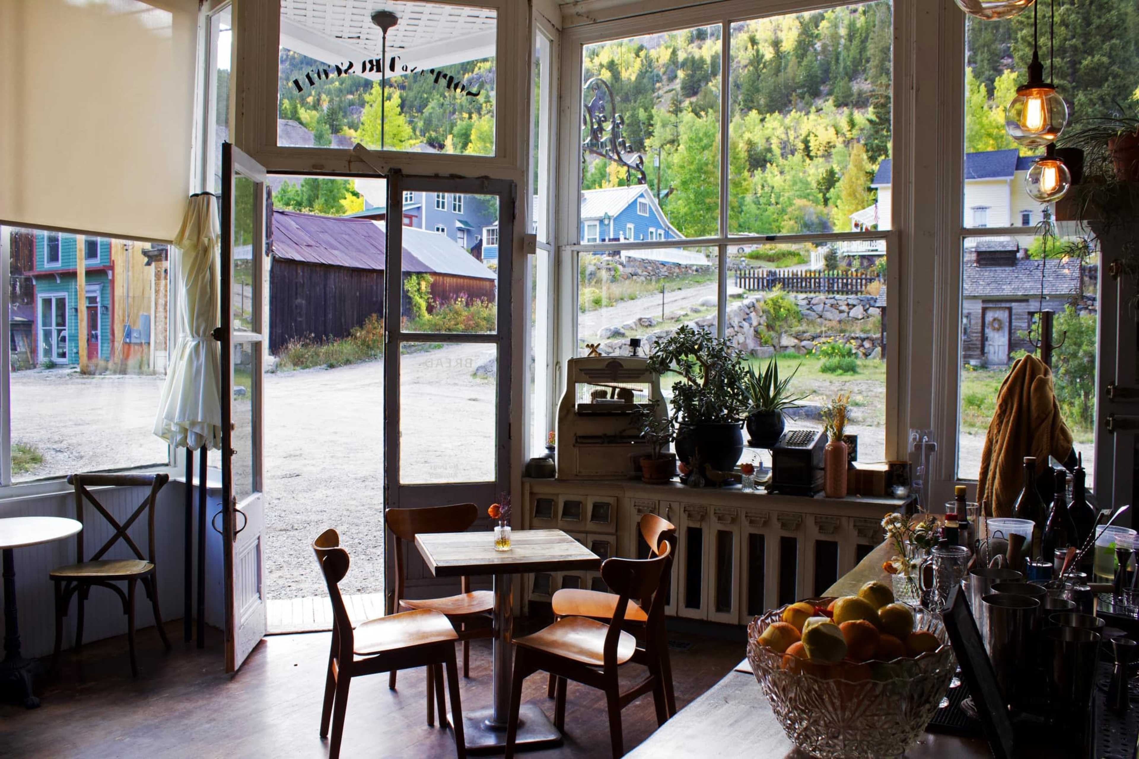 The image shows a bright café interior with wooden tables and chairs, large windows overlooking a scenic outdoor landscape.