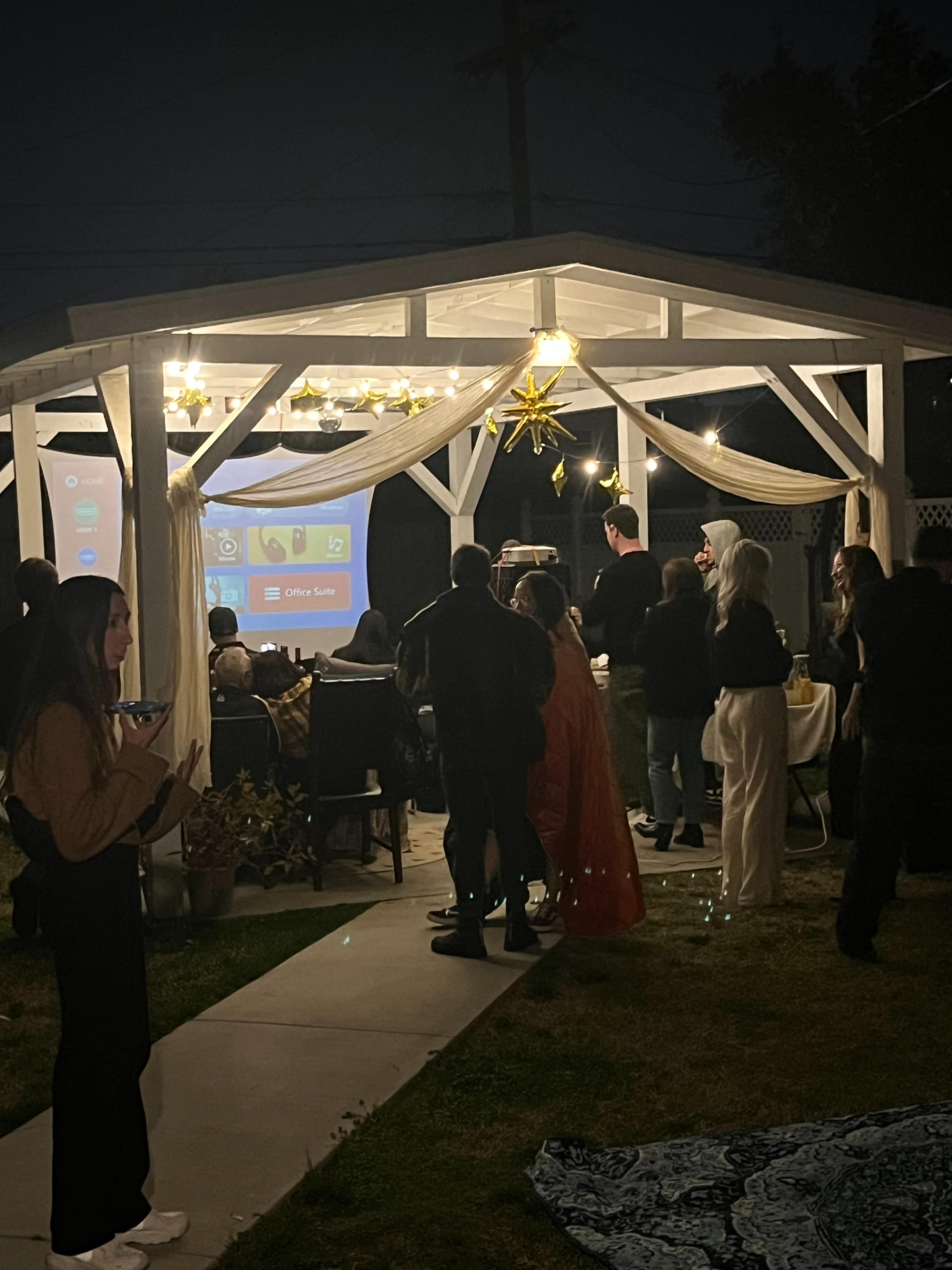 A group of people gathers under a lit outdoor structure to watch a presentation projected onto a screen during an evening event.