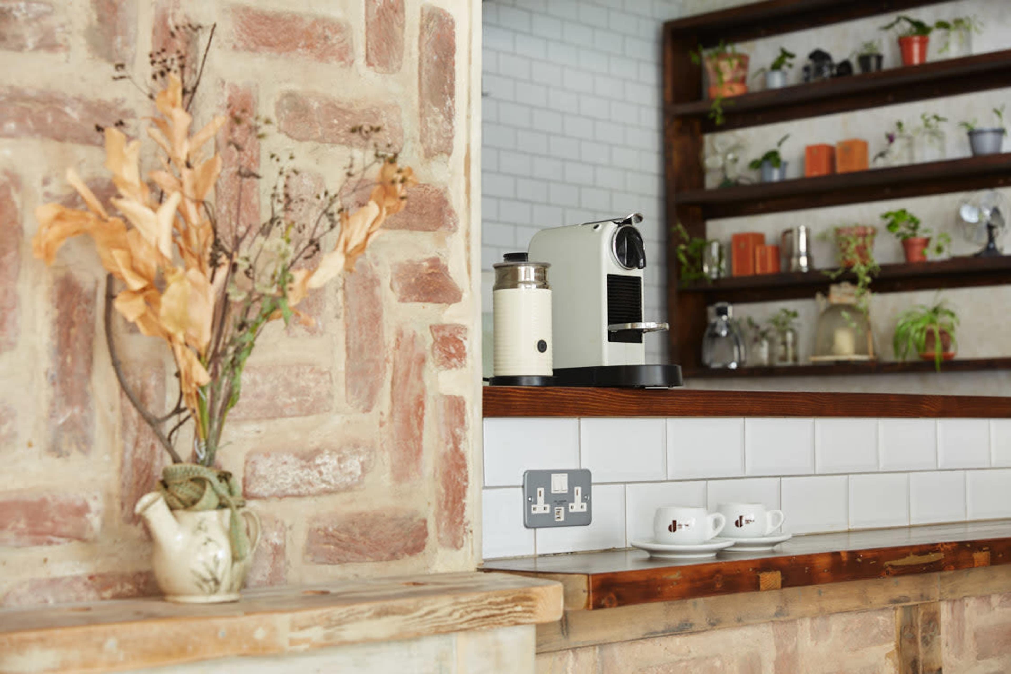 A kitchen with a coffee machine on a wooden countertop, a vase of dried flowers, and shelves containing plants and decorative items.