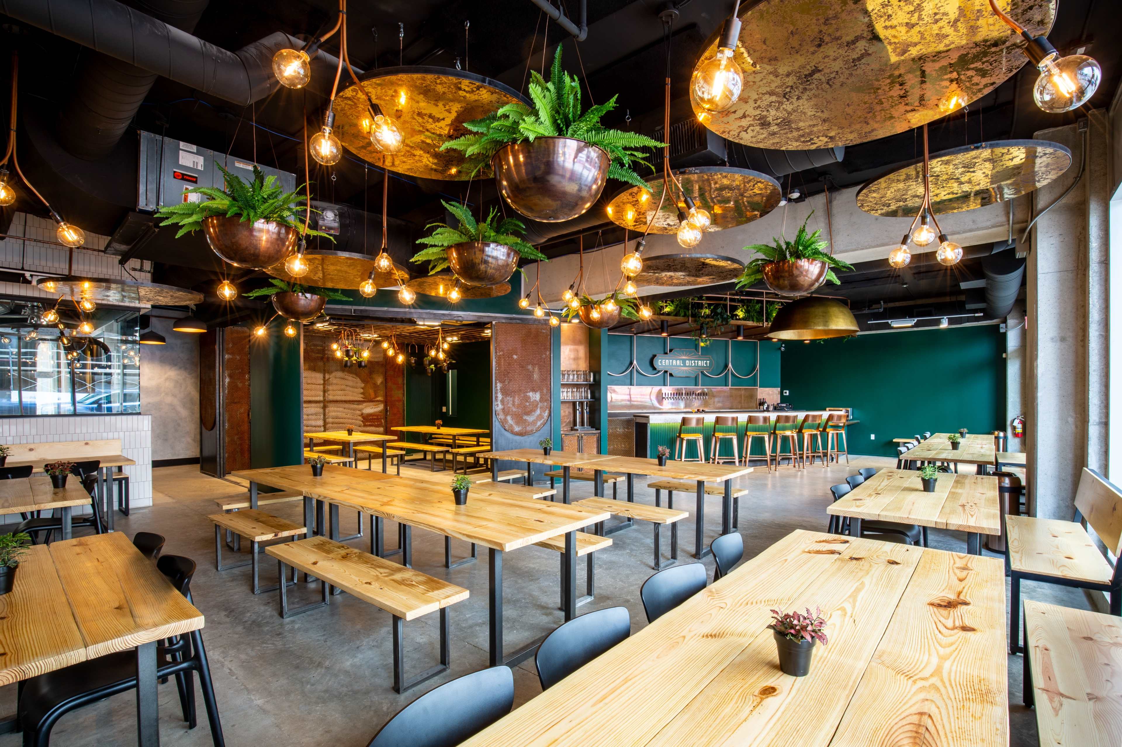 A modern restaurant interior features long wooden tables, black chairs, and hanging light fixtures with greenery against a backdrop of teal and metallic decor.
