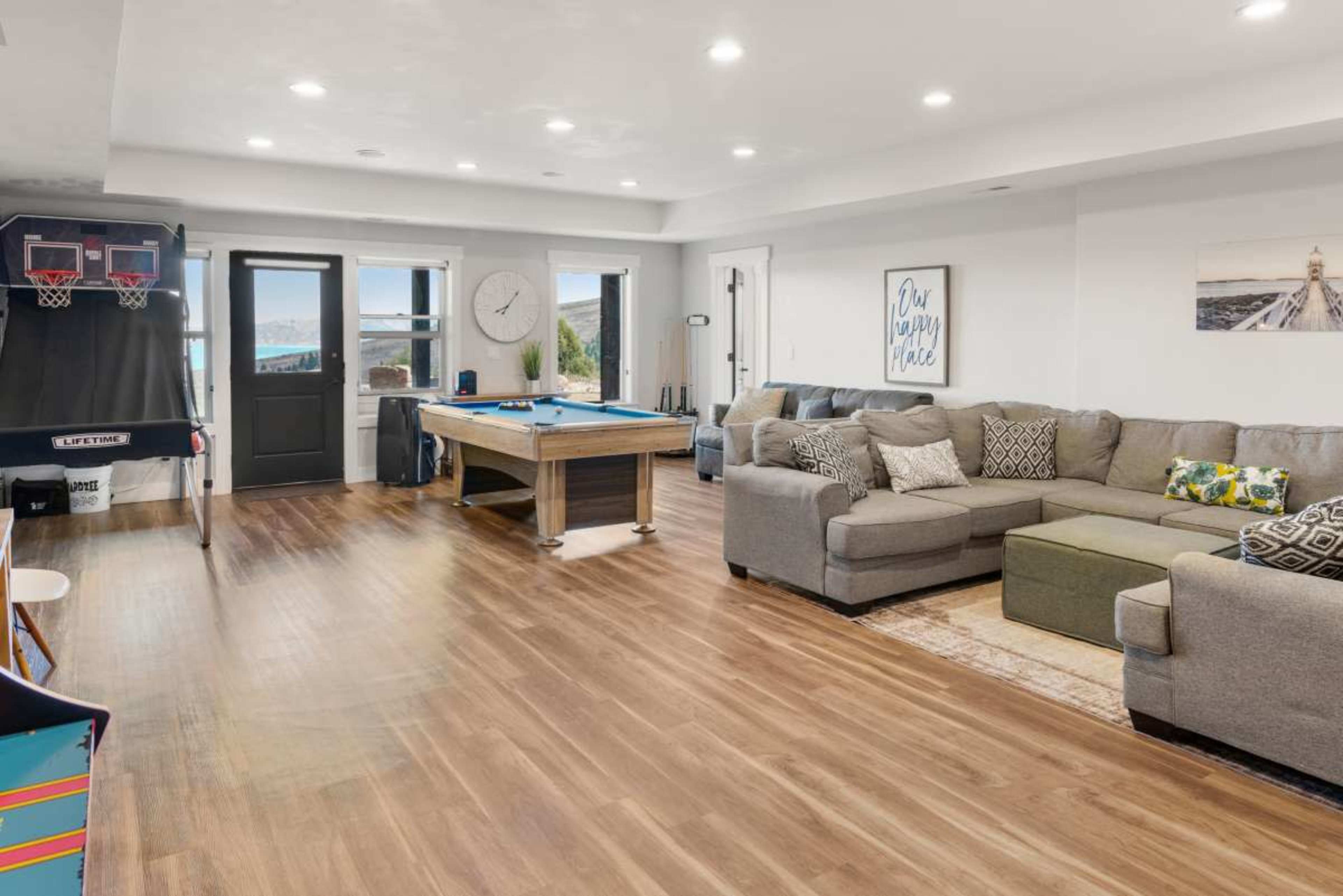 The image shows a spacious living area featuring a sectional sofa, a pool table, and a small basketball arcade game, with large windows providing natural light.