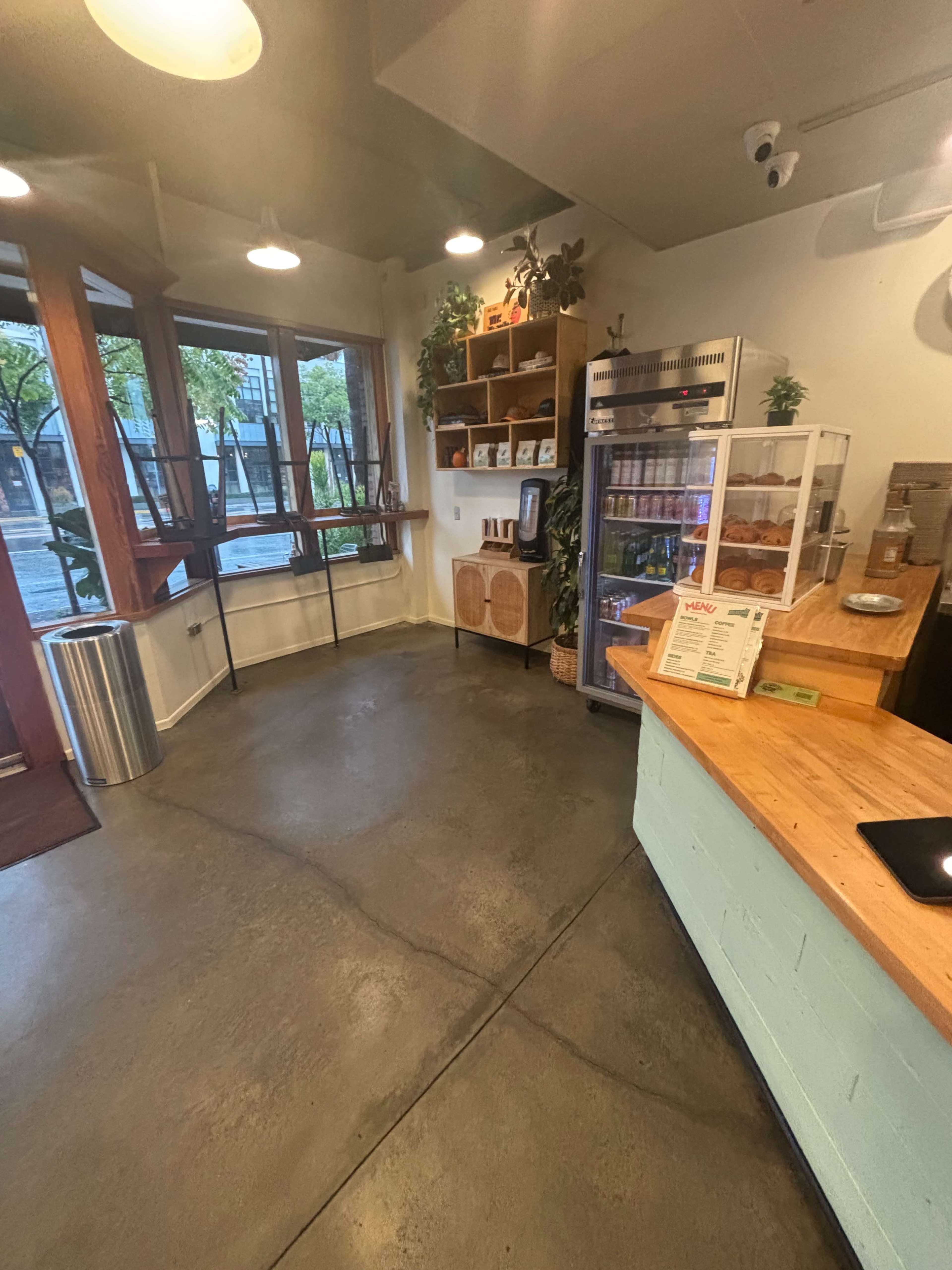 The image shows the interior of a small café with a wooden counter, display case, shelves filled with products, and large windows facing an outdoor area.