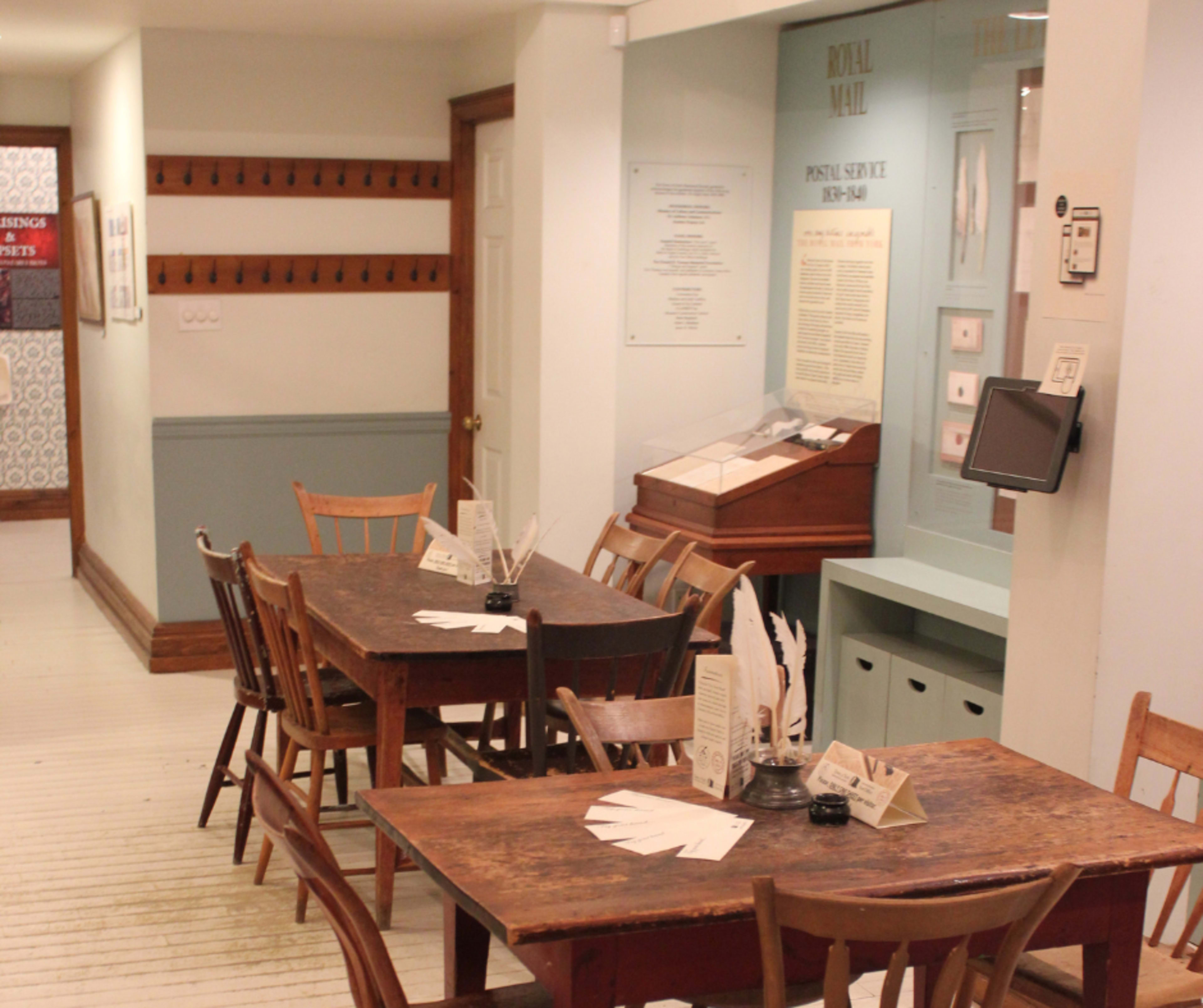 The image shows a small, vintage-style cafe or dining area with wooden tables and chairs, along with informational displays and a postal service exhibit on the walls.