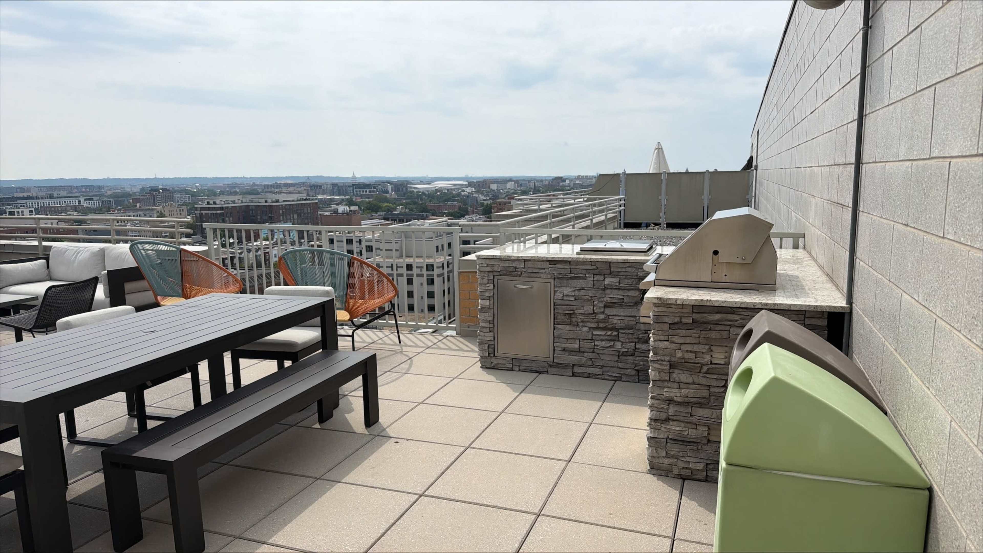 The image shows a rooftop terrace with outdoor furniture, a stone barbecue area, and a view of the city skyline.