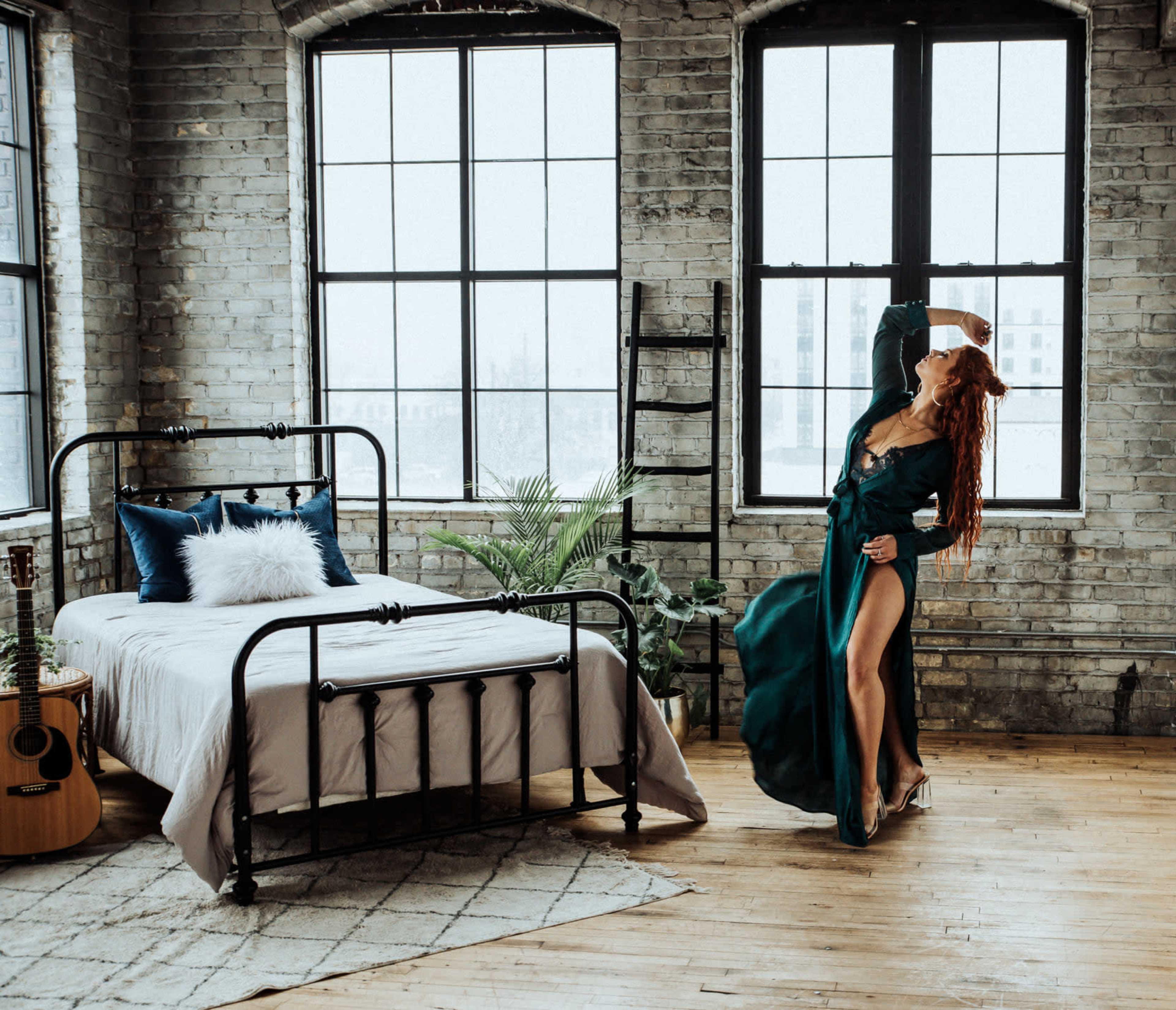 A person in a green dress poses near a large window in an industrial-style bedroom with a bed, guitar, and plants.
