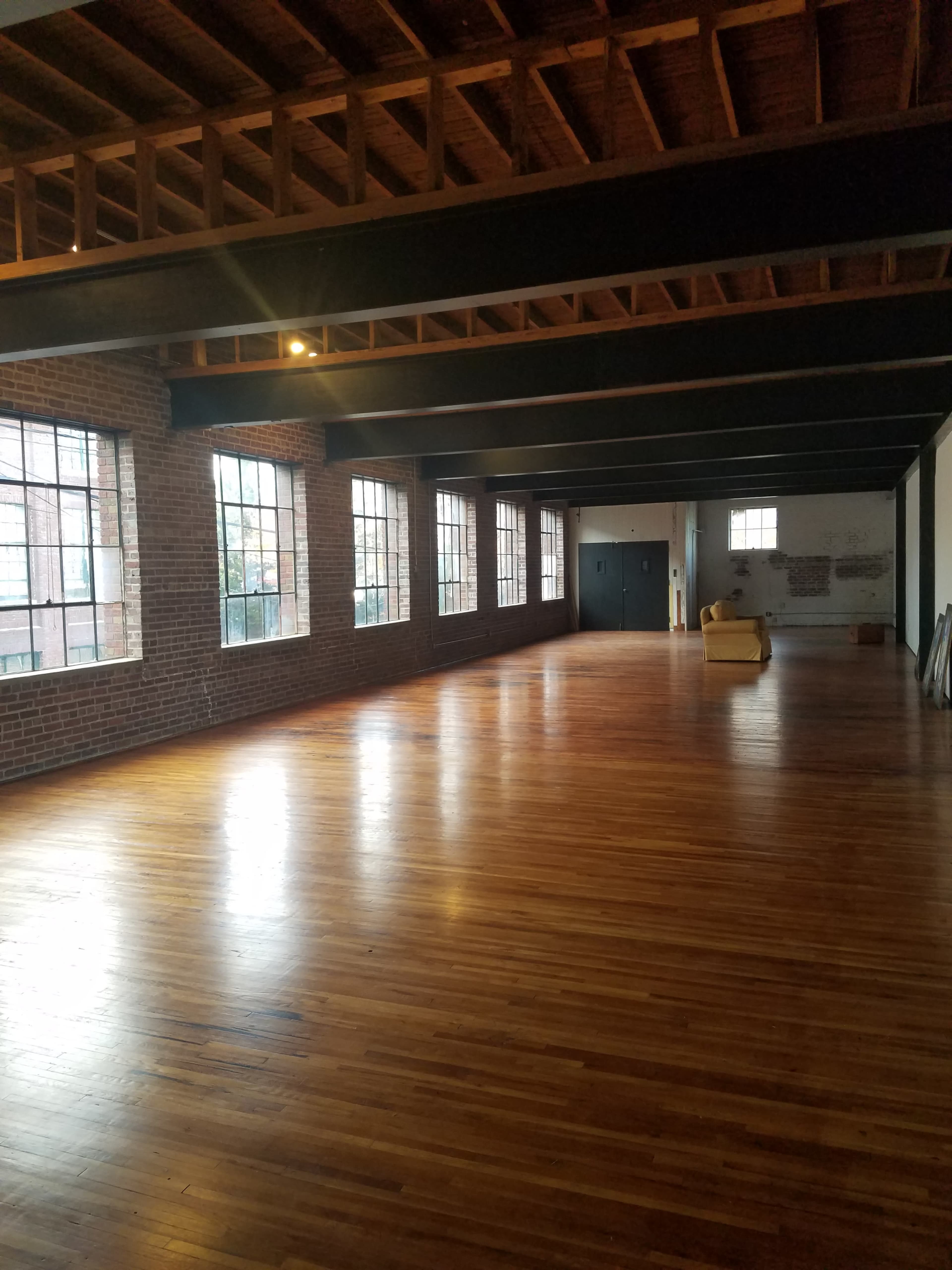 A spacious, empty room with large windows, exposed brick walls, and wooden flooring beneath a beam ceiling.