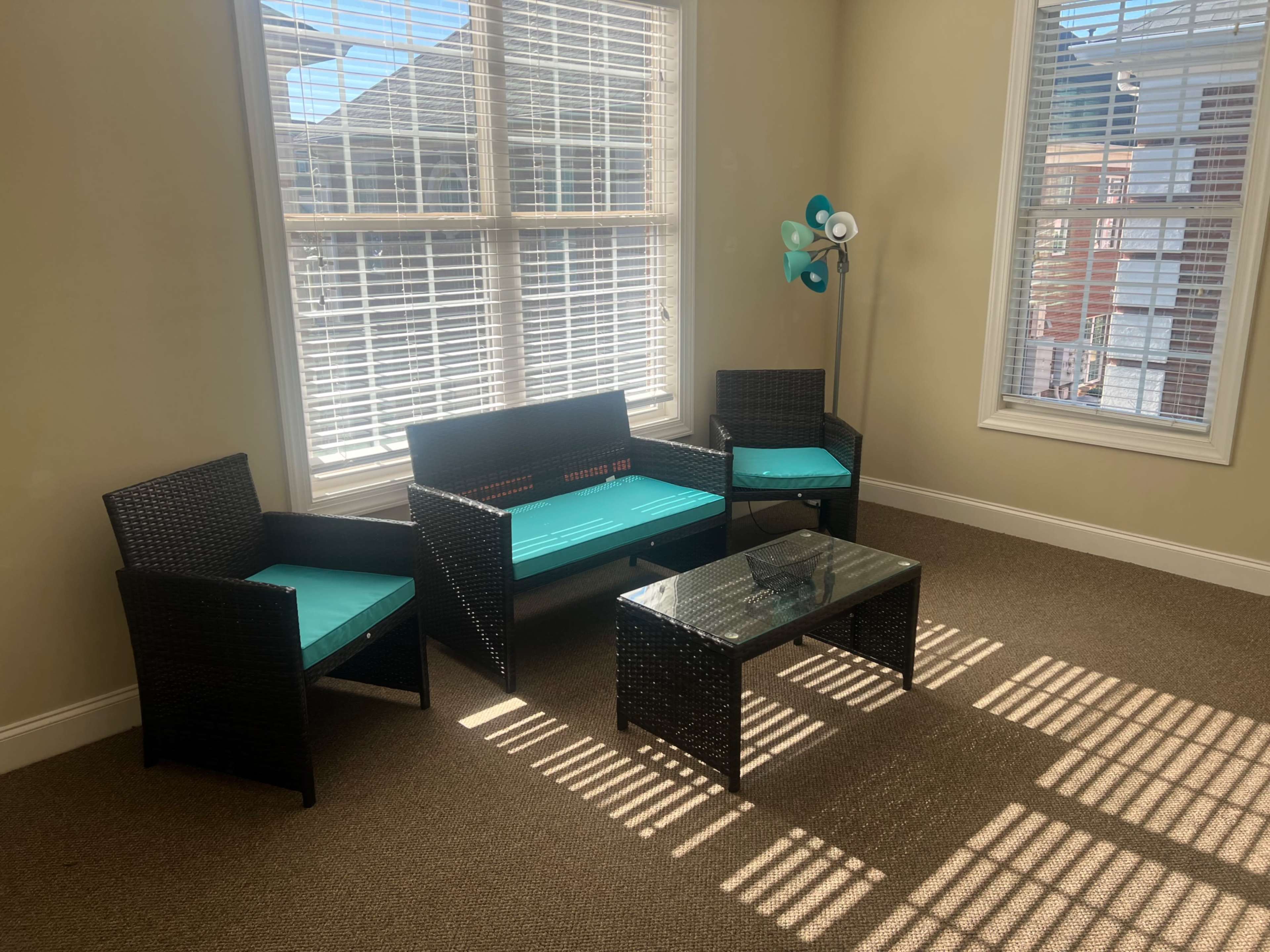 The image shows a seating area with a black wicker sofa and two chairs, accented with turquoise cushions, and a glass coffee table, all positioned under large windows that let in sunlight.
