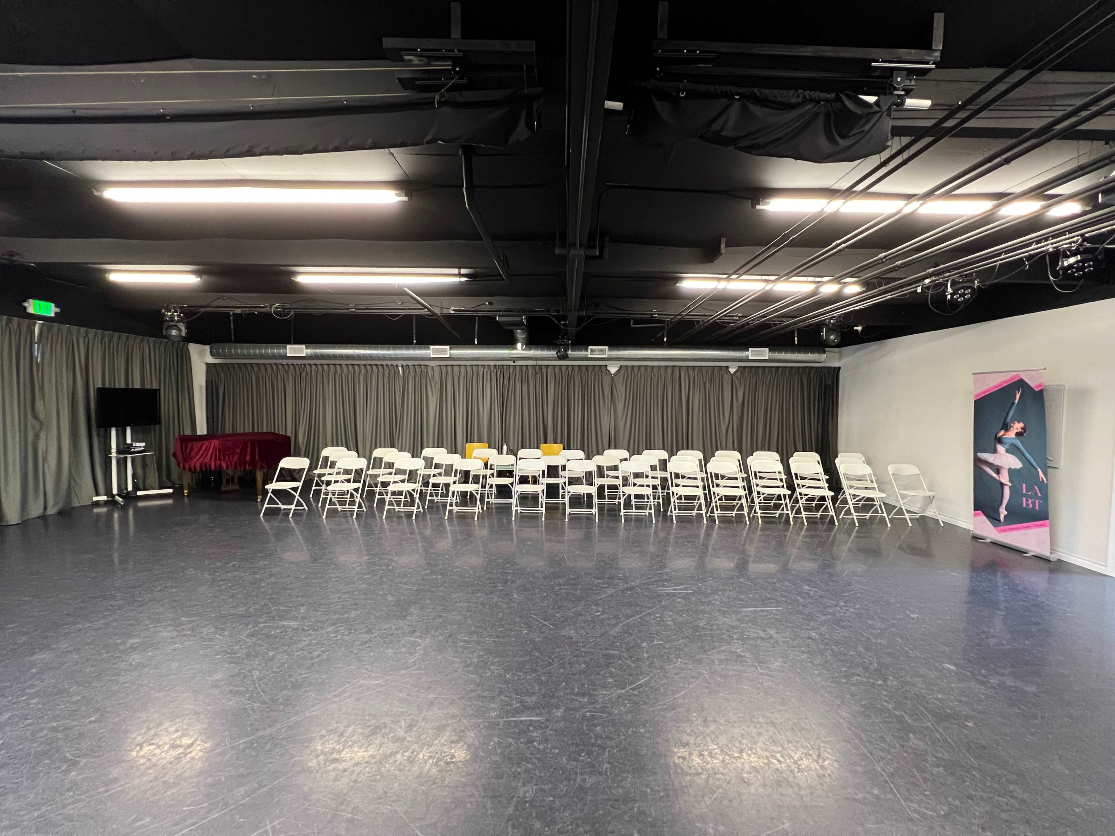 The image shows a dance studio with a row of white chairs set up facing a stage area, flanked by curtains and a banner.
