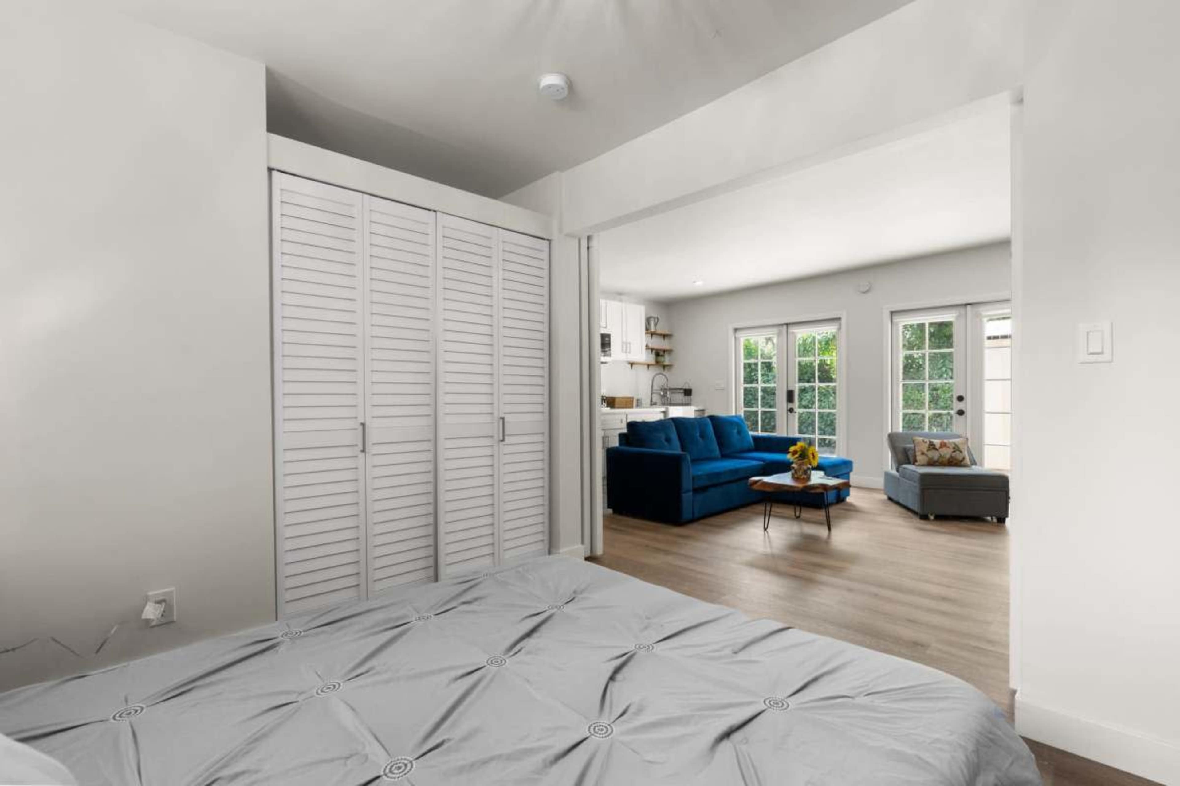 The image shows a bedroom with a bed in the foreground and a living area visible through an open doorway, featuring a blue couch and a coffee table.