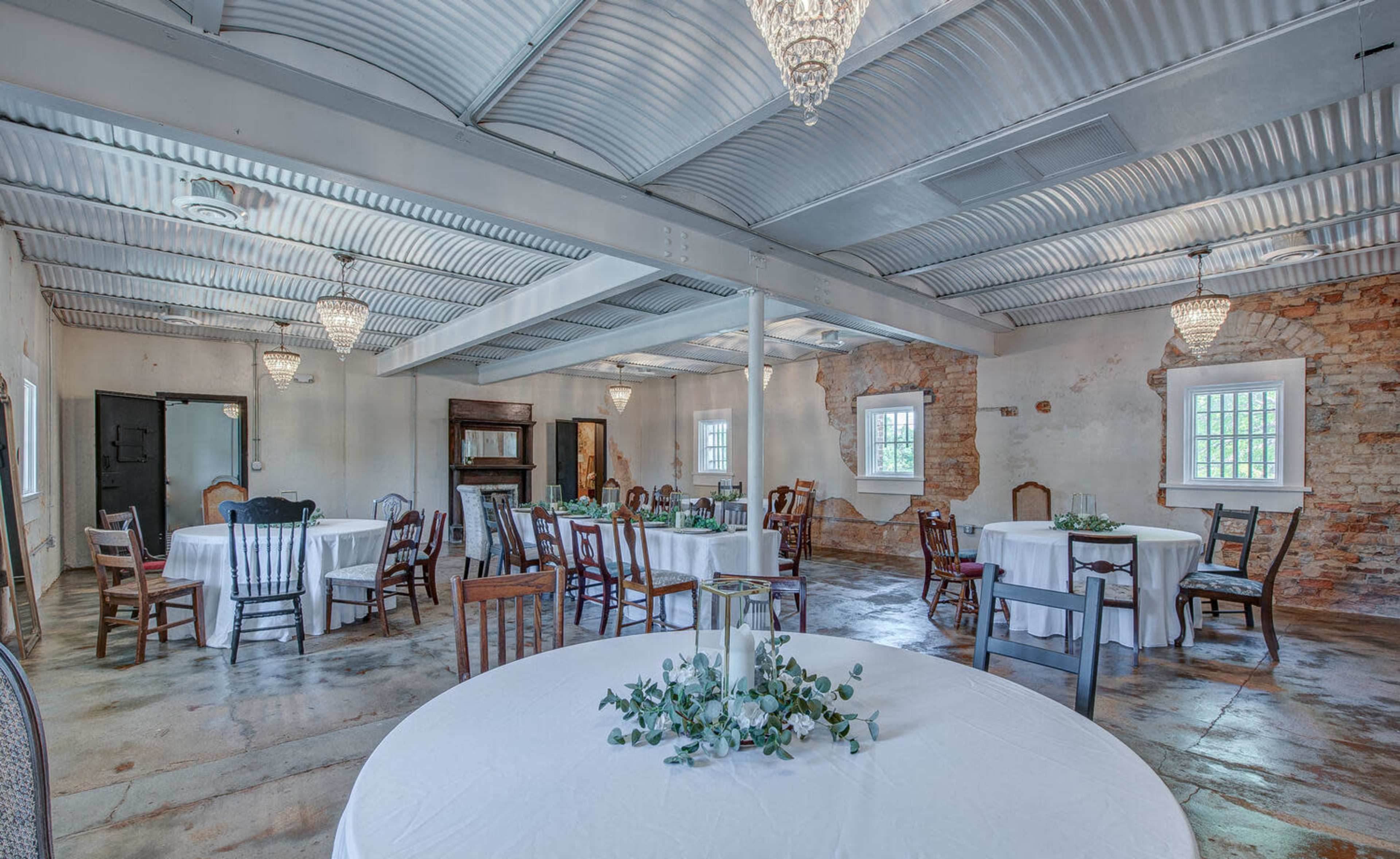 A spacious event room with round tables covered in white linens, surrounded by a mix of wooden chairs, and featuring exposed brick walls and chandeliers.