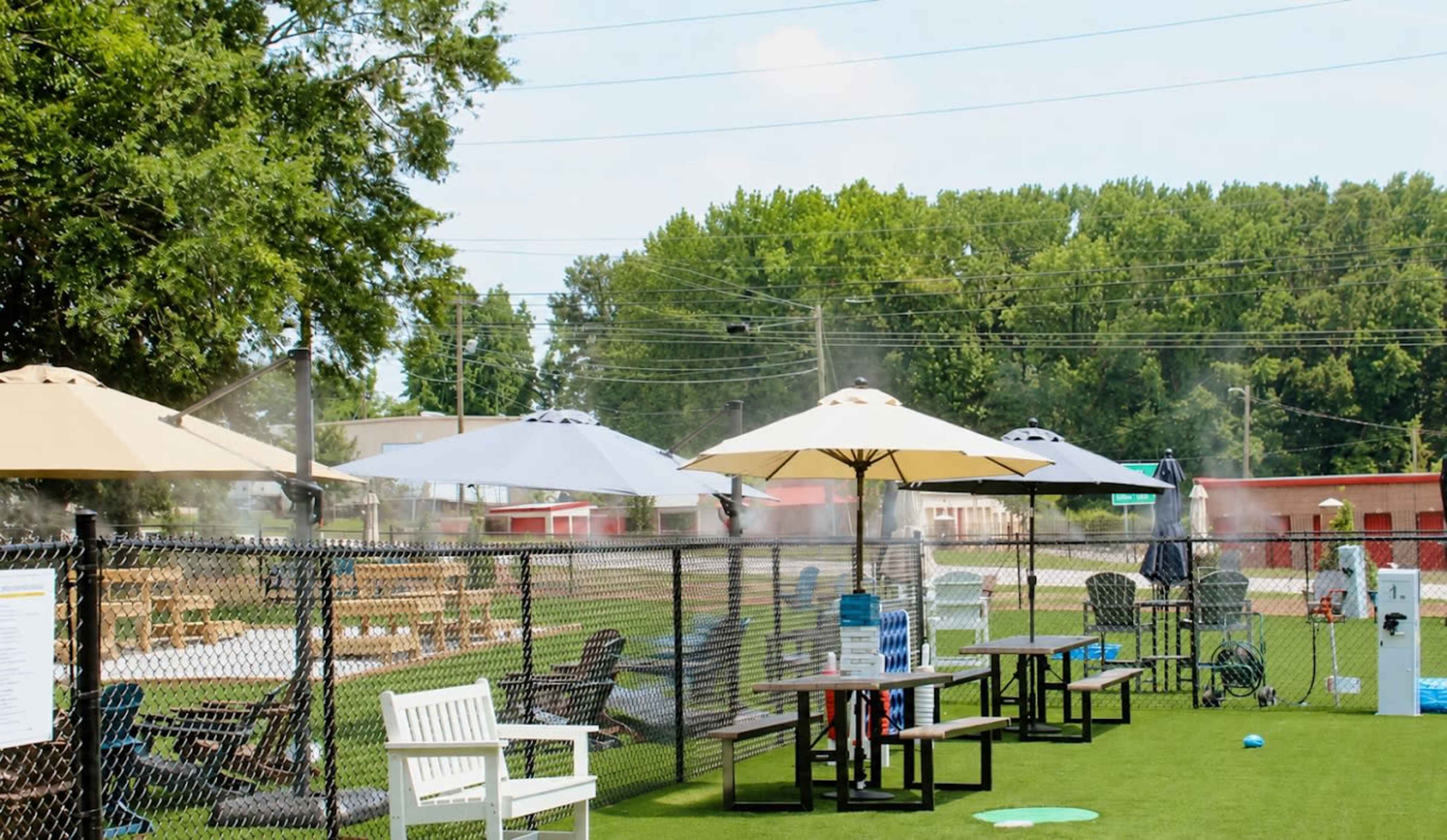 The image shows an outdoor recreational area with misting umbrellas, seating arrangements, and a fenced grassy space.