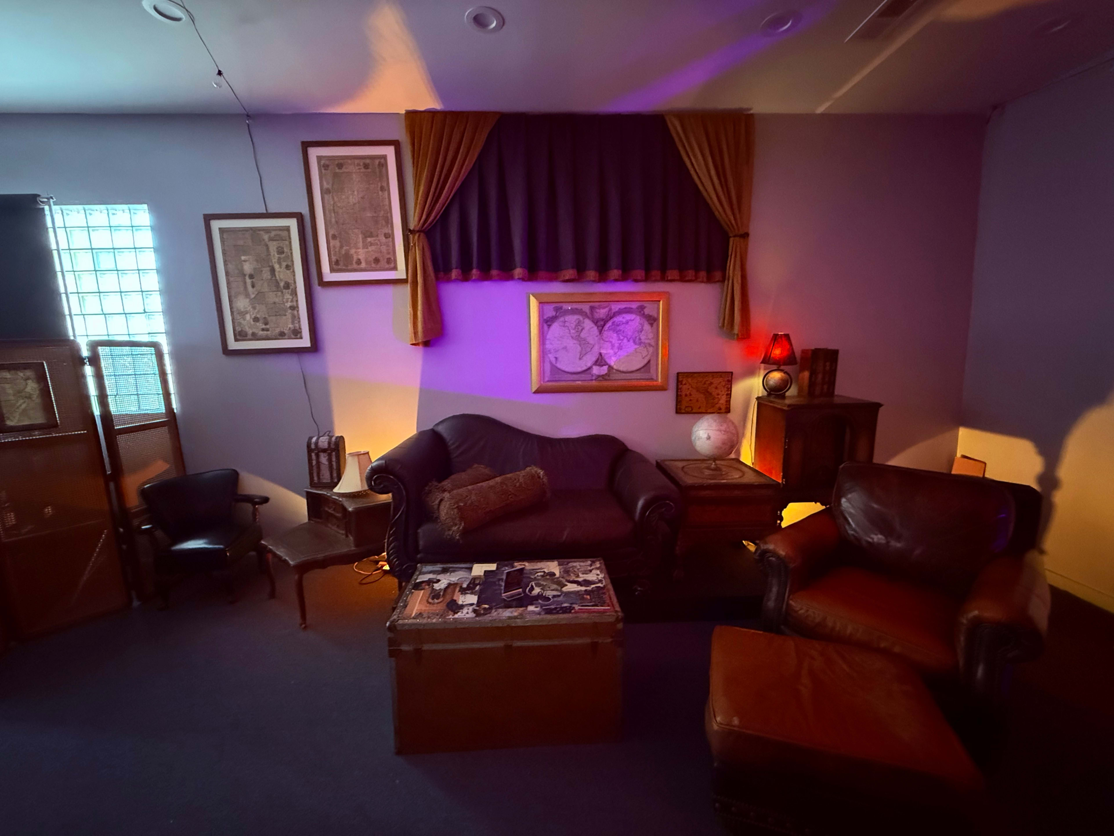 The image shows a cozy room with a dark blue wall, featuring a mix of vintage furniture including a couch, two armchairs, a coffee table, and various framed artworks.