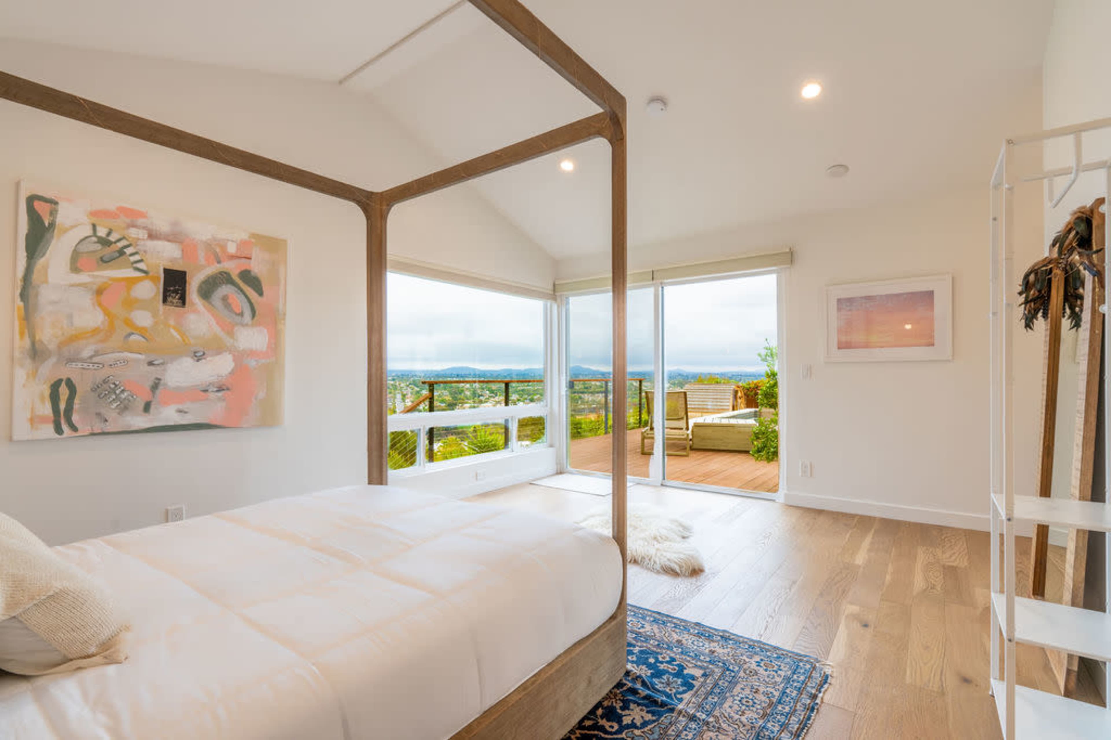 A spacious bedroom features a four-poster bed, large windows showcasing a view of the outdoors, and modern decor with artwork on the walls.