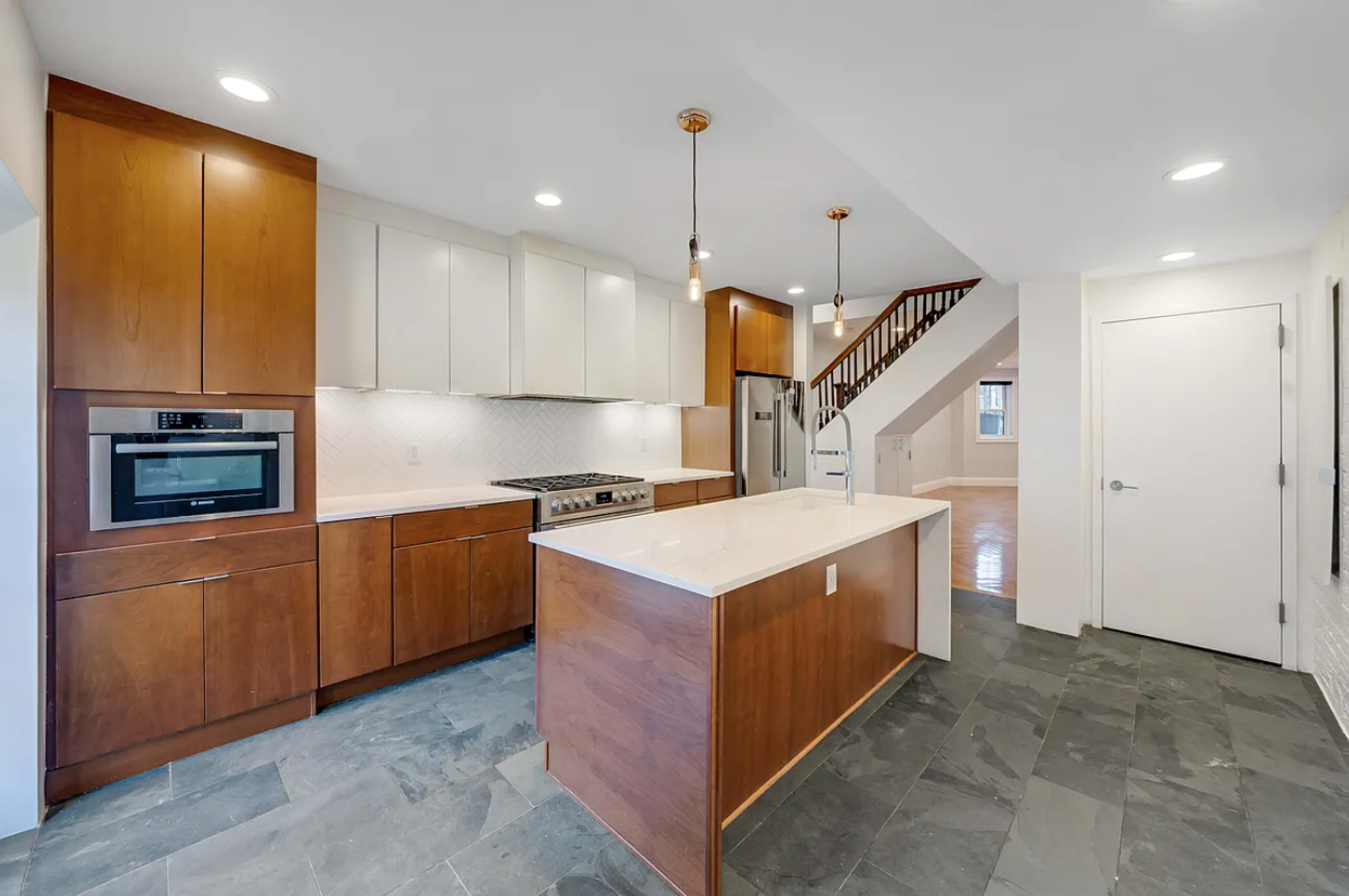 The image shows a modern kitchen featuring wooden cabinets, a large island, stainless steel appliances, and a staircase in the background.