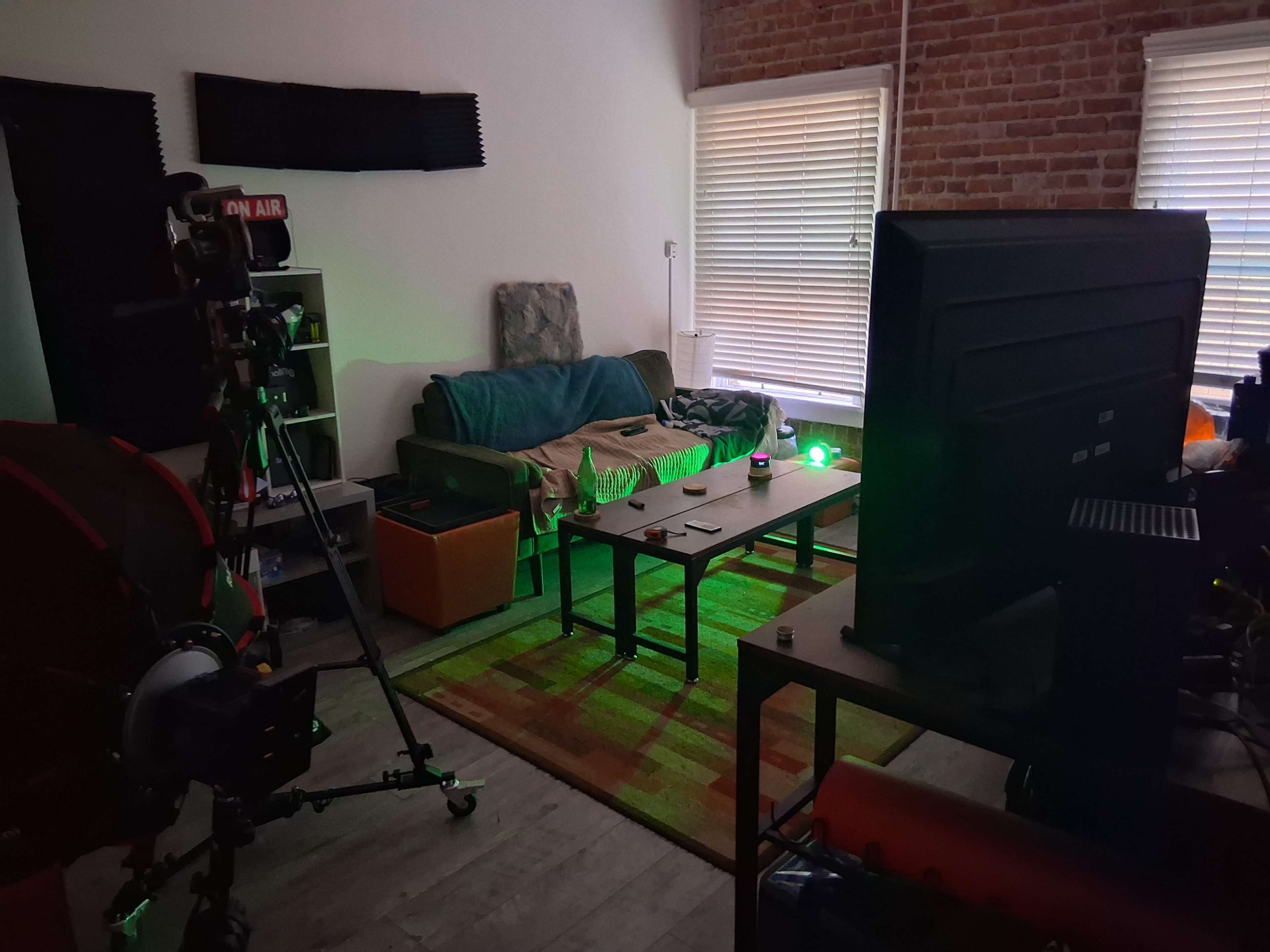 The image shows a dimly lit room with a couch, coffee table, and various electronic equipment, including a camera and a large monitor.