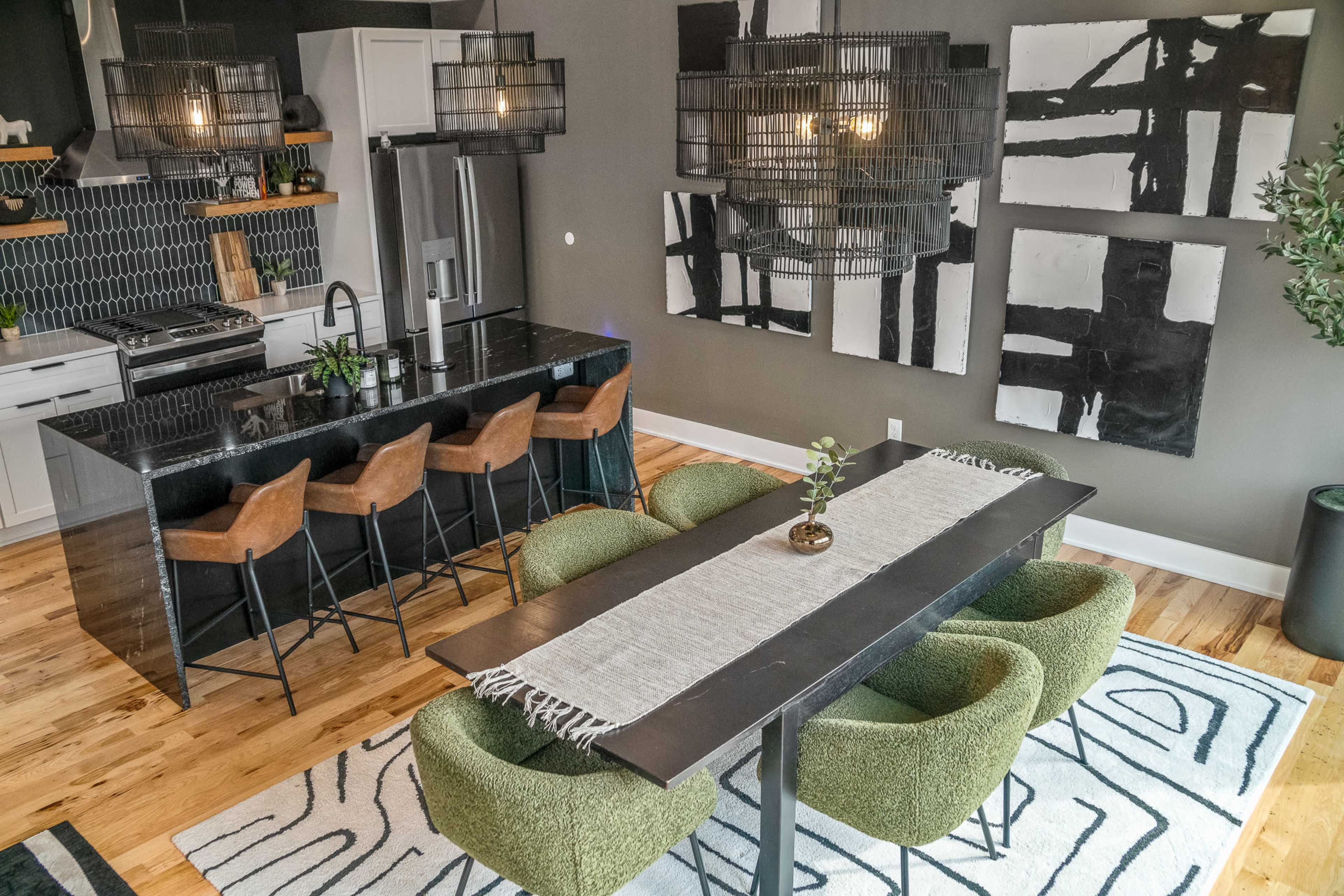 A modern dining area features a long black table surrounded by green chairs, with a bar and kitchen visible in the background, complemented by pendant lights and abstract wall art.