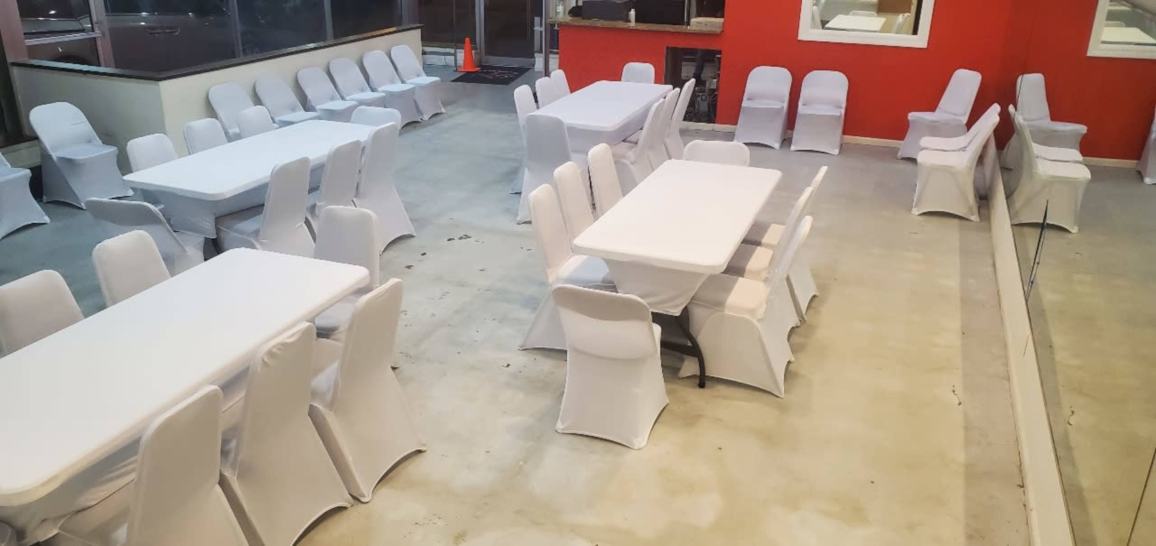 The image shows a large indoor space with multiple tables and chairs covered in white fabric, arranged neatly on a concrete floor.
