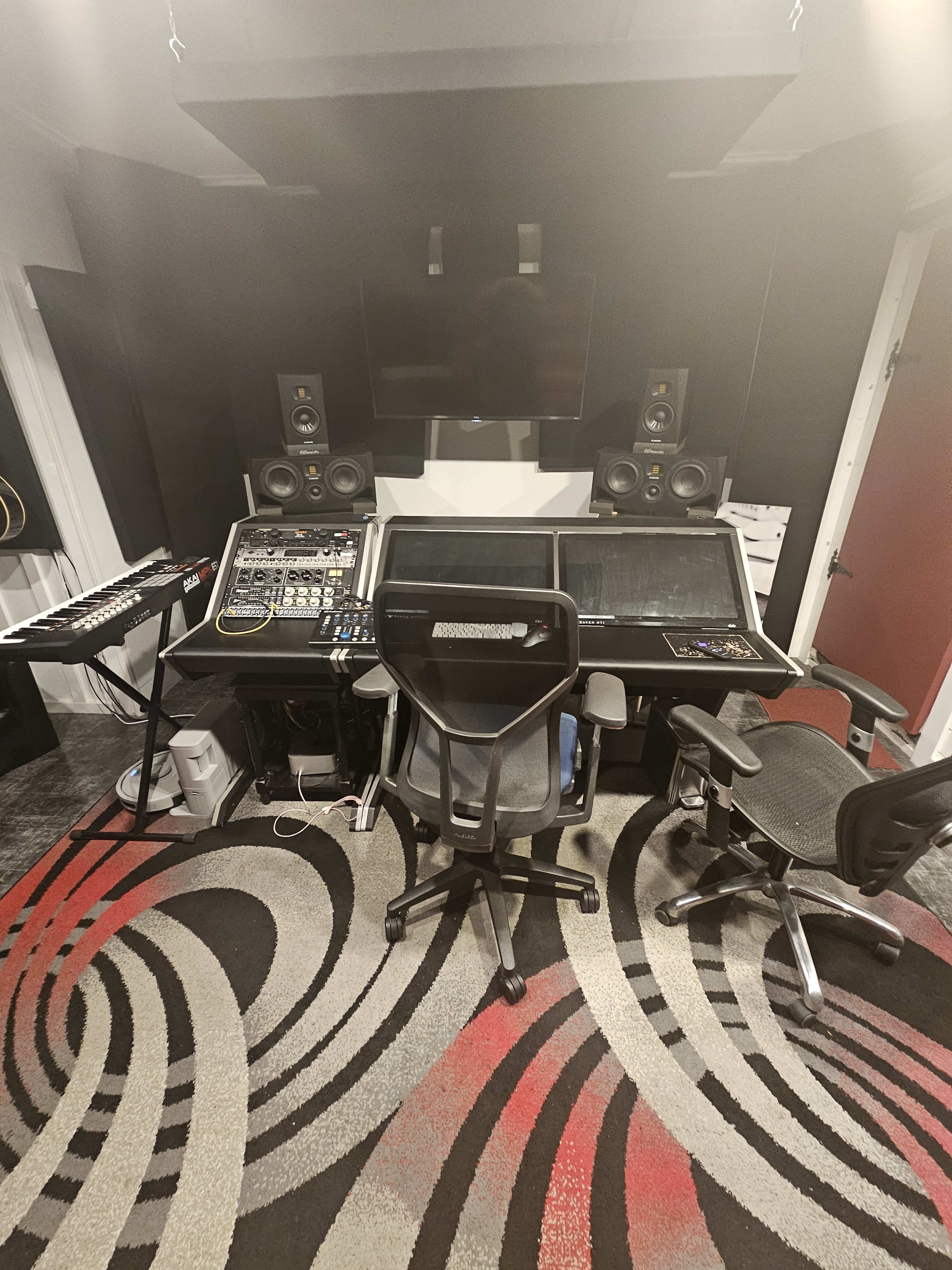 The image shows a music recording studio featuring a large mixing console, two speaker monitors, keyboard, and three office chairs arranged on a patterned carpet.