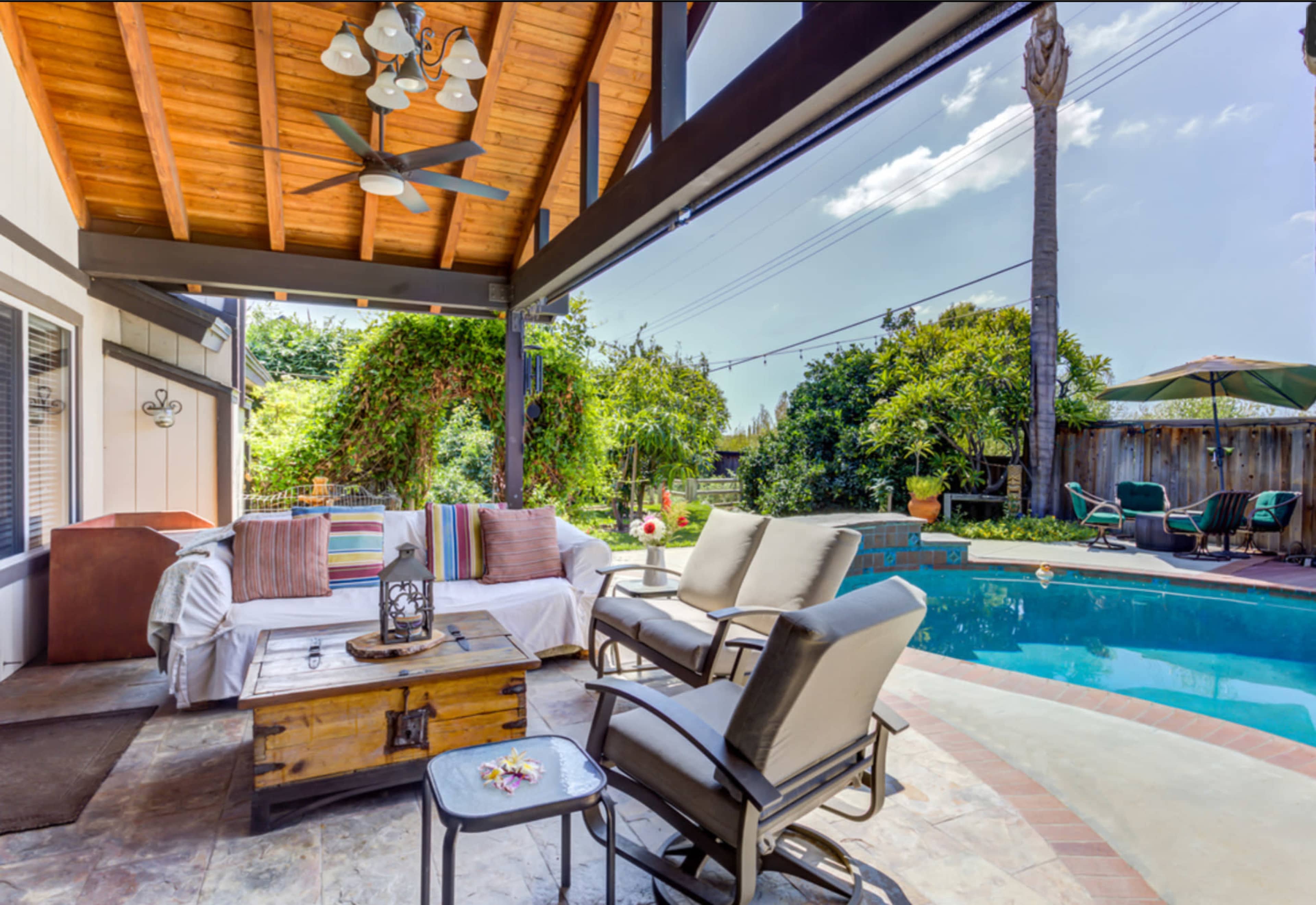 A covered patio features a seating area with a coffee table and a view of a swimming pool surrounded by greenery.