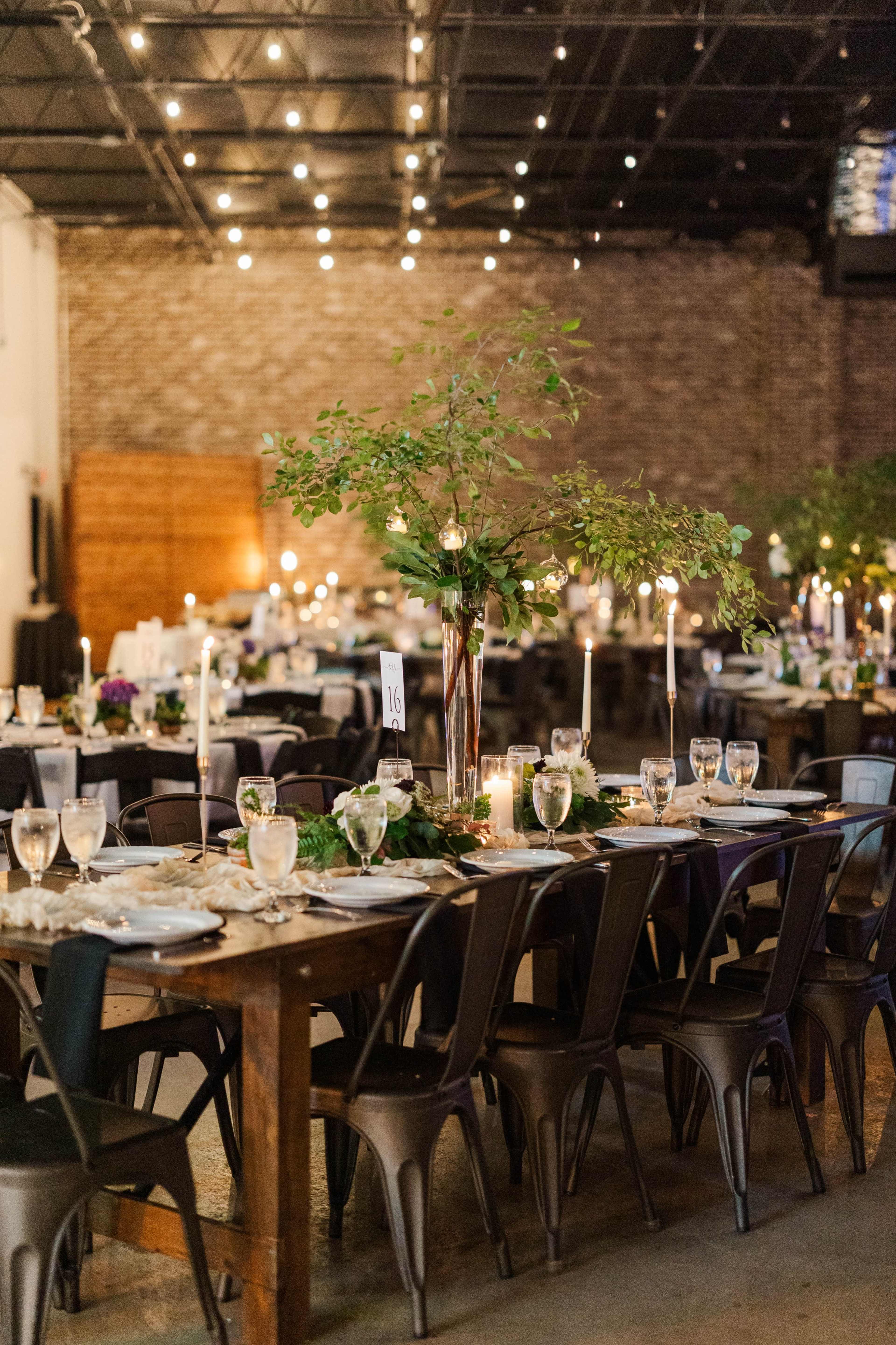 The image shows a decorated dining area with long wooden tables set with glassware, candles, and floral centerpieces in an industrial space with string lights overhead.