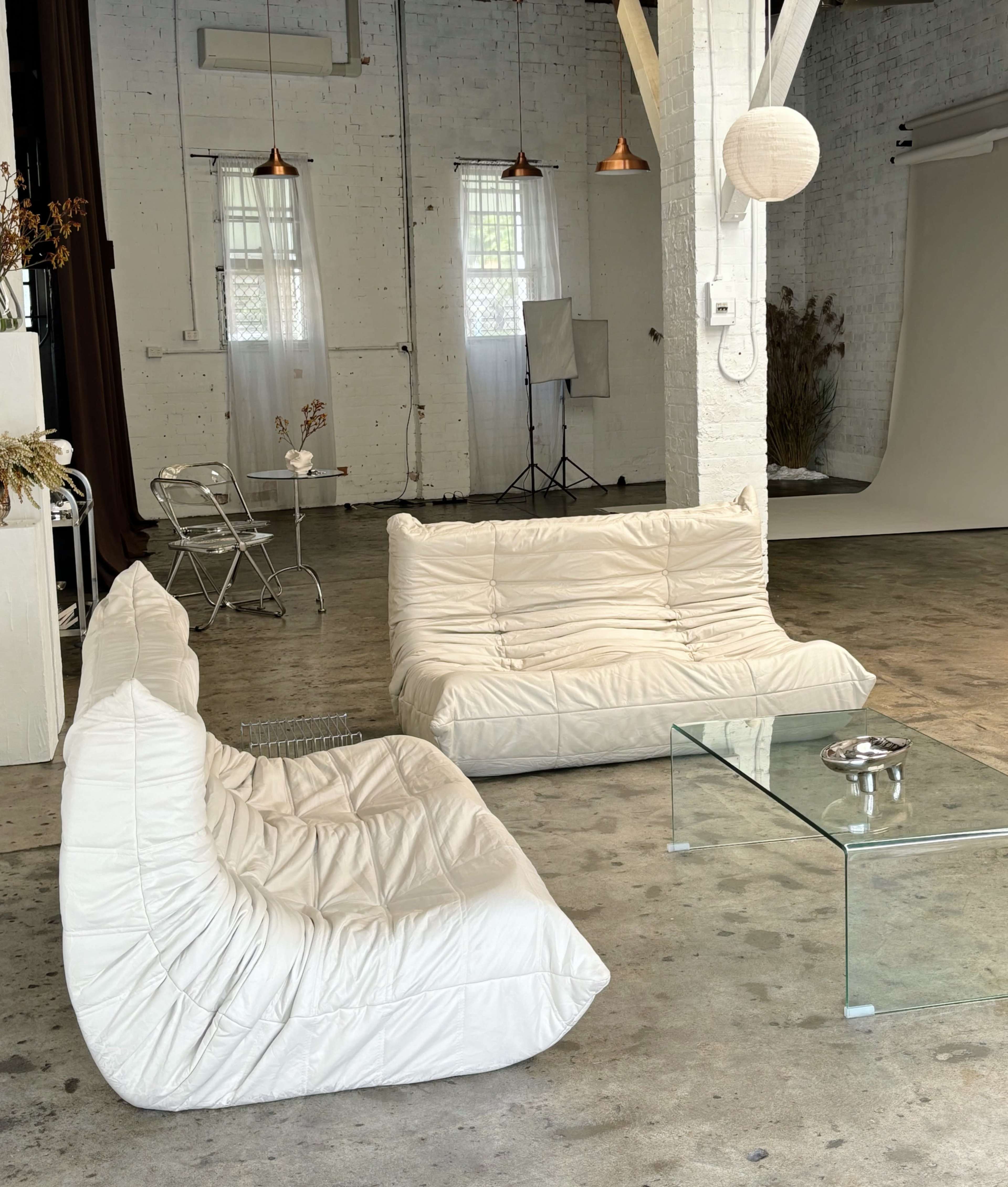 The scene features two white modular sofas and a glass coffee table in a spacious, industrial-style room with exposed brick walls and wood beams.