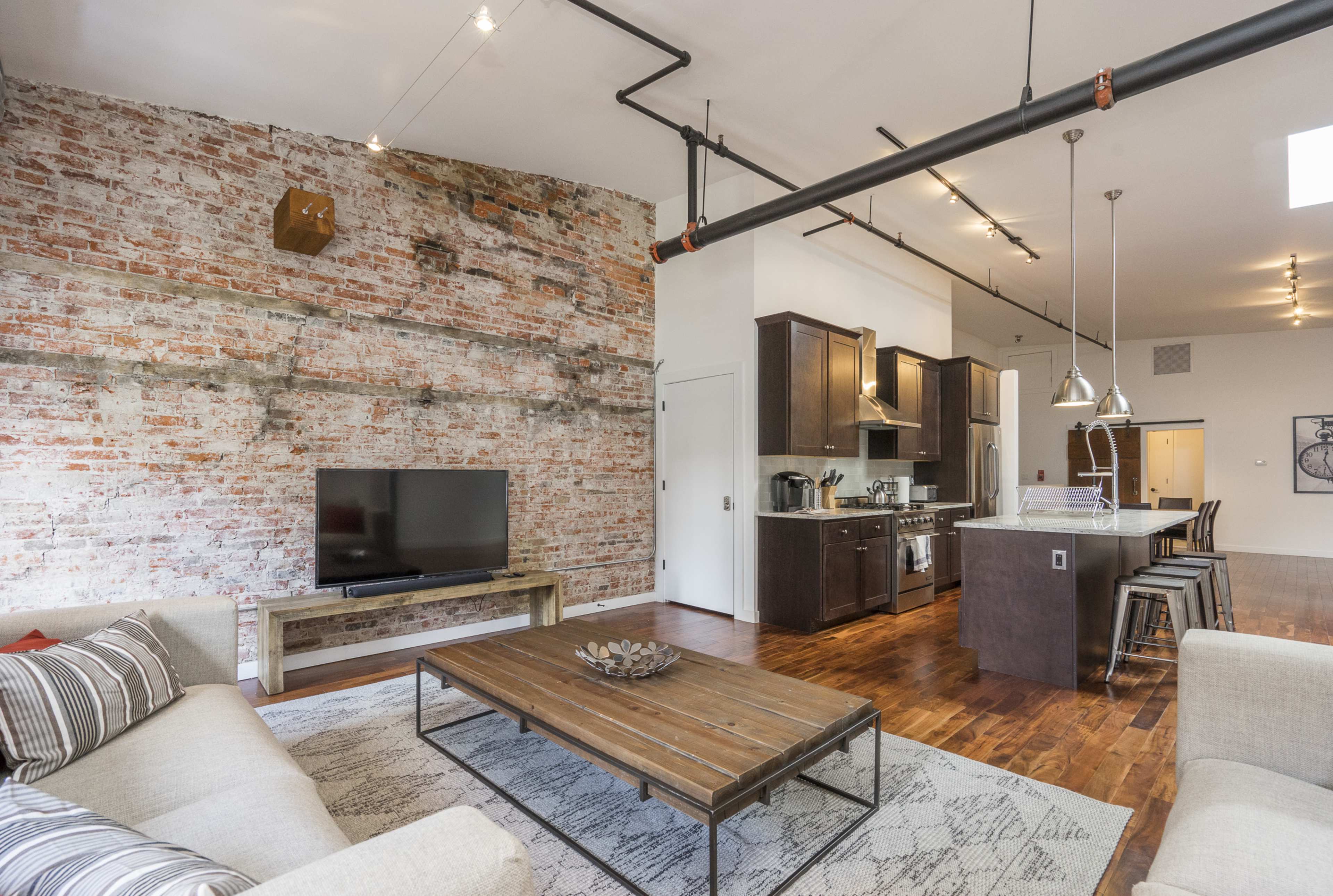 The image shows an open-concept living area with a brick accent wall, a modern kitchen, and a comfortable seating area featuring a large coffee table and a television.