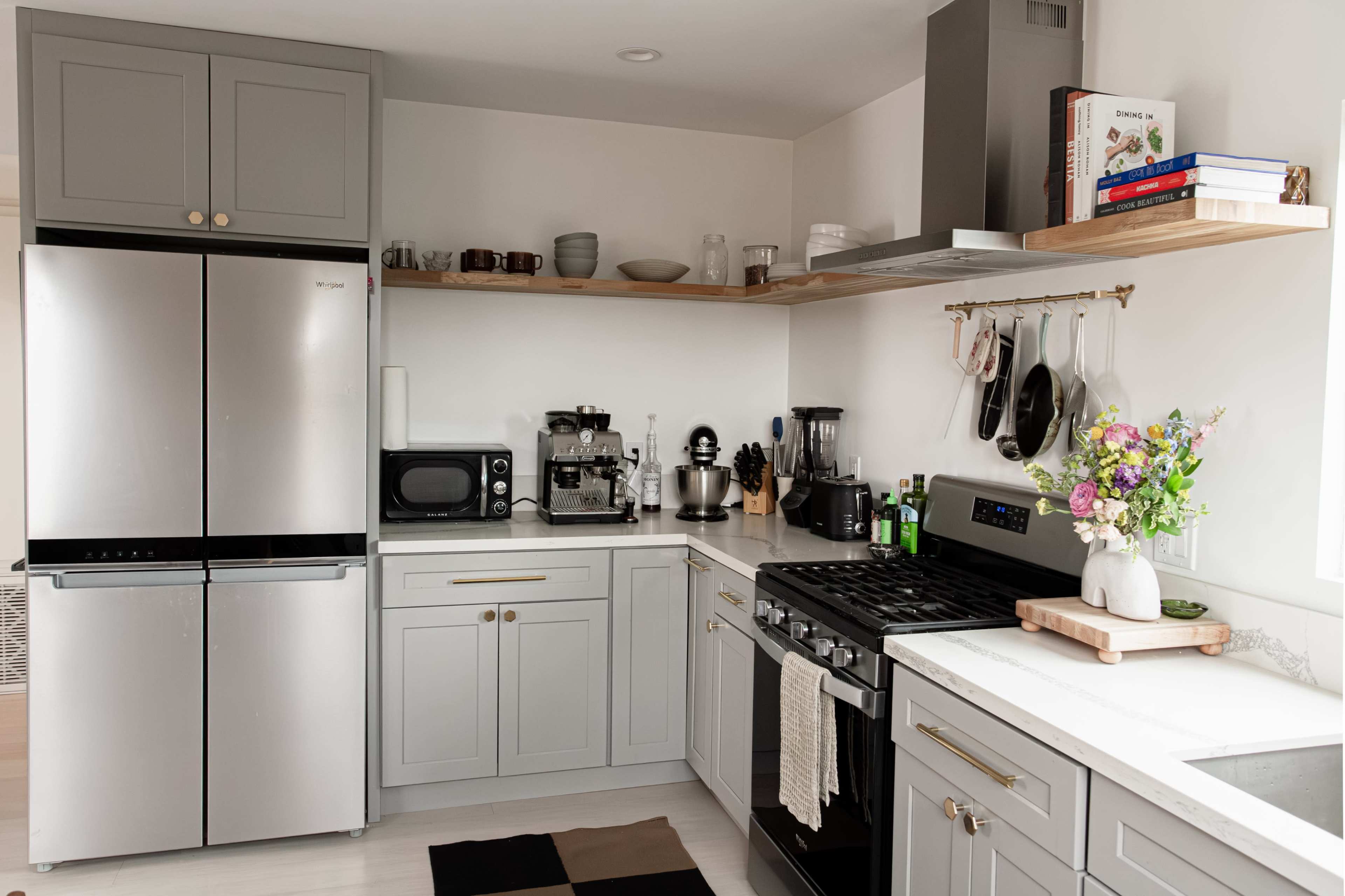 A modern kitchen features stainless steel appliances, light gray cabinetry, open shelving, and a countertop with a small vase of flowers.