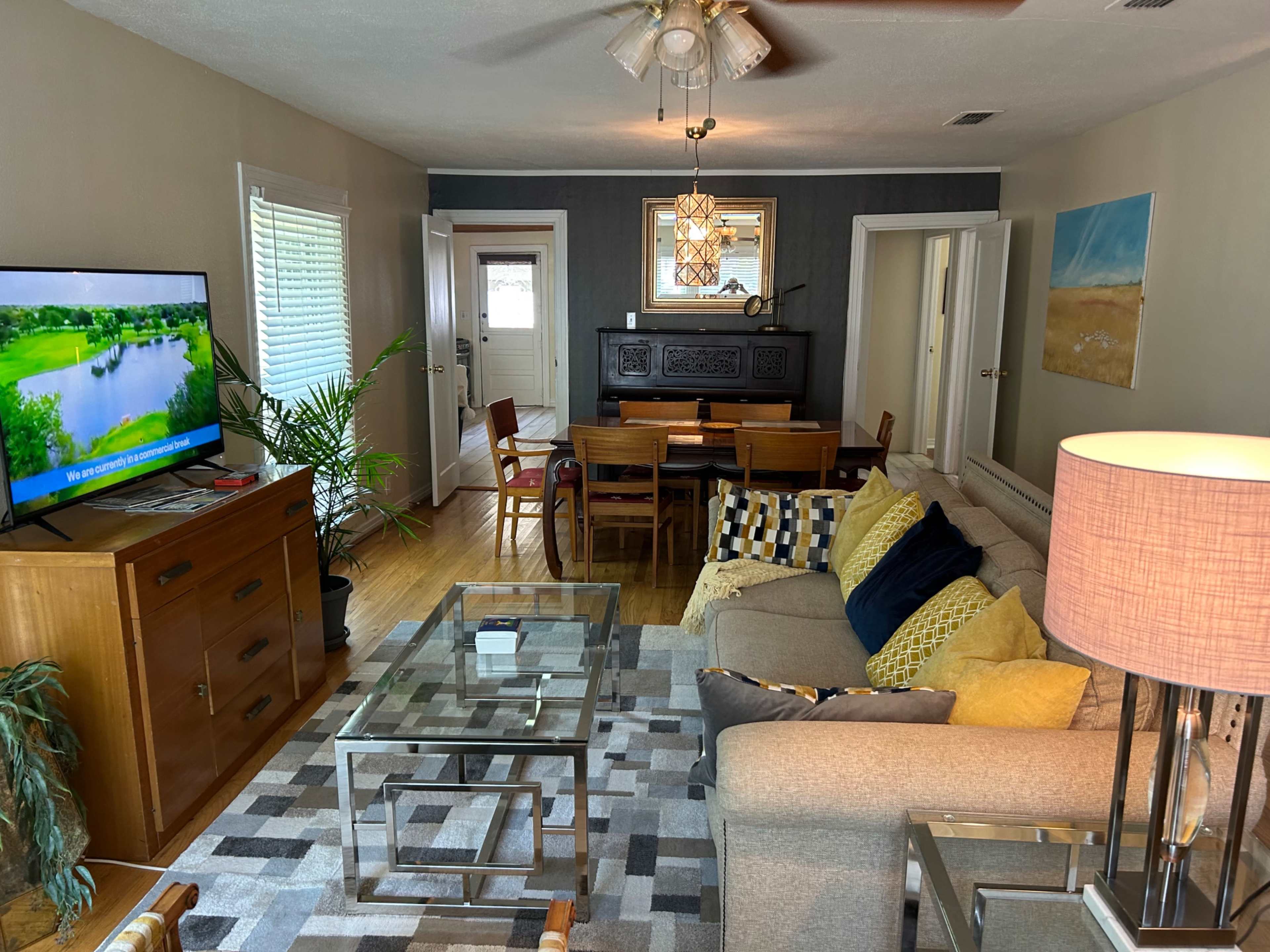 The living room features a gray sofa with colorful pillows, a glass coffee table, a wooden sideboard, and a dining area with wooden chairs, complemented by a large television displaying a landscape scene.