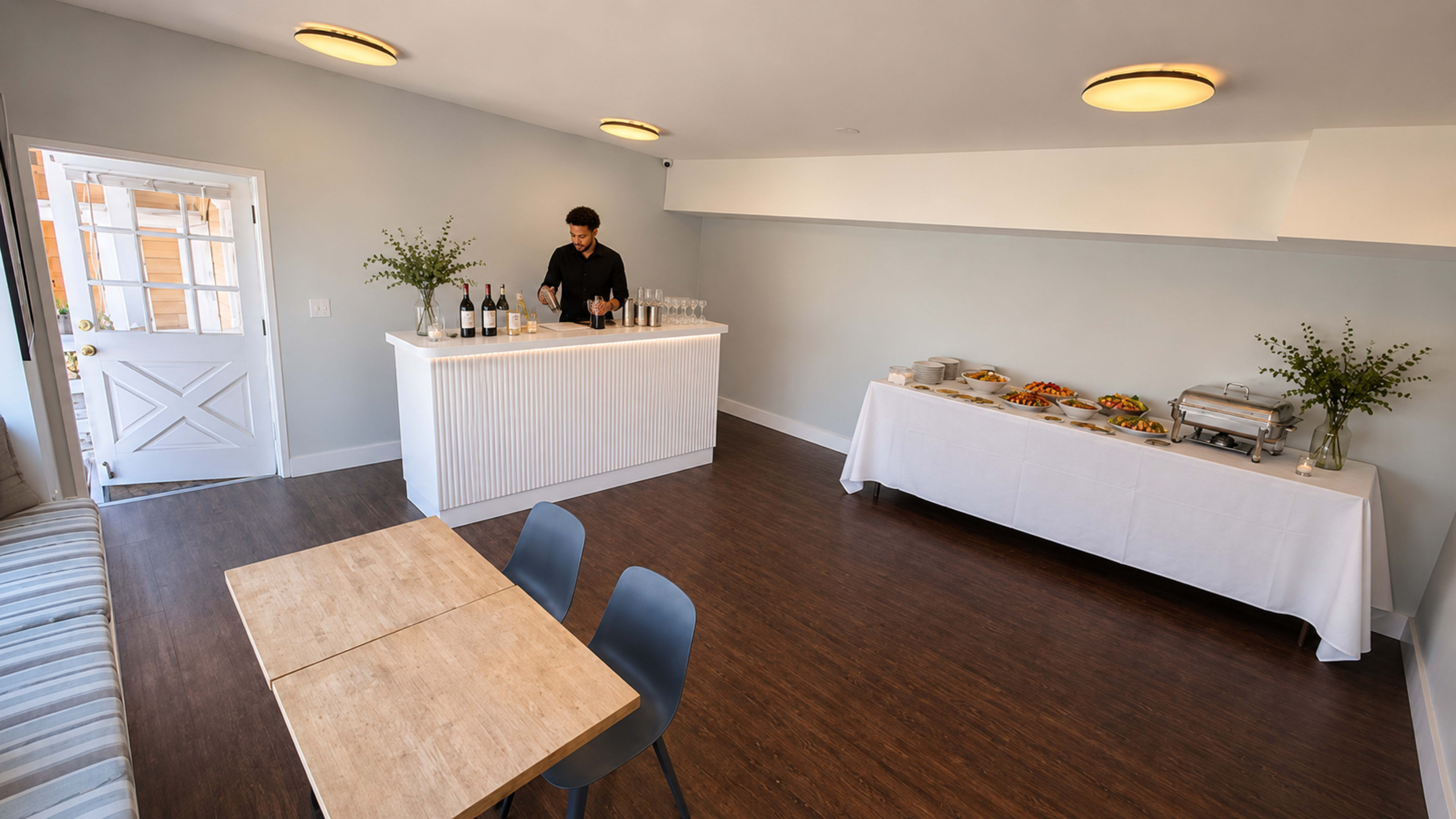 A bartender prepares drinks at a counter in a minimalist event space with a table of food set against a light-colored wall.