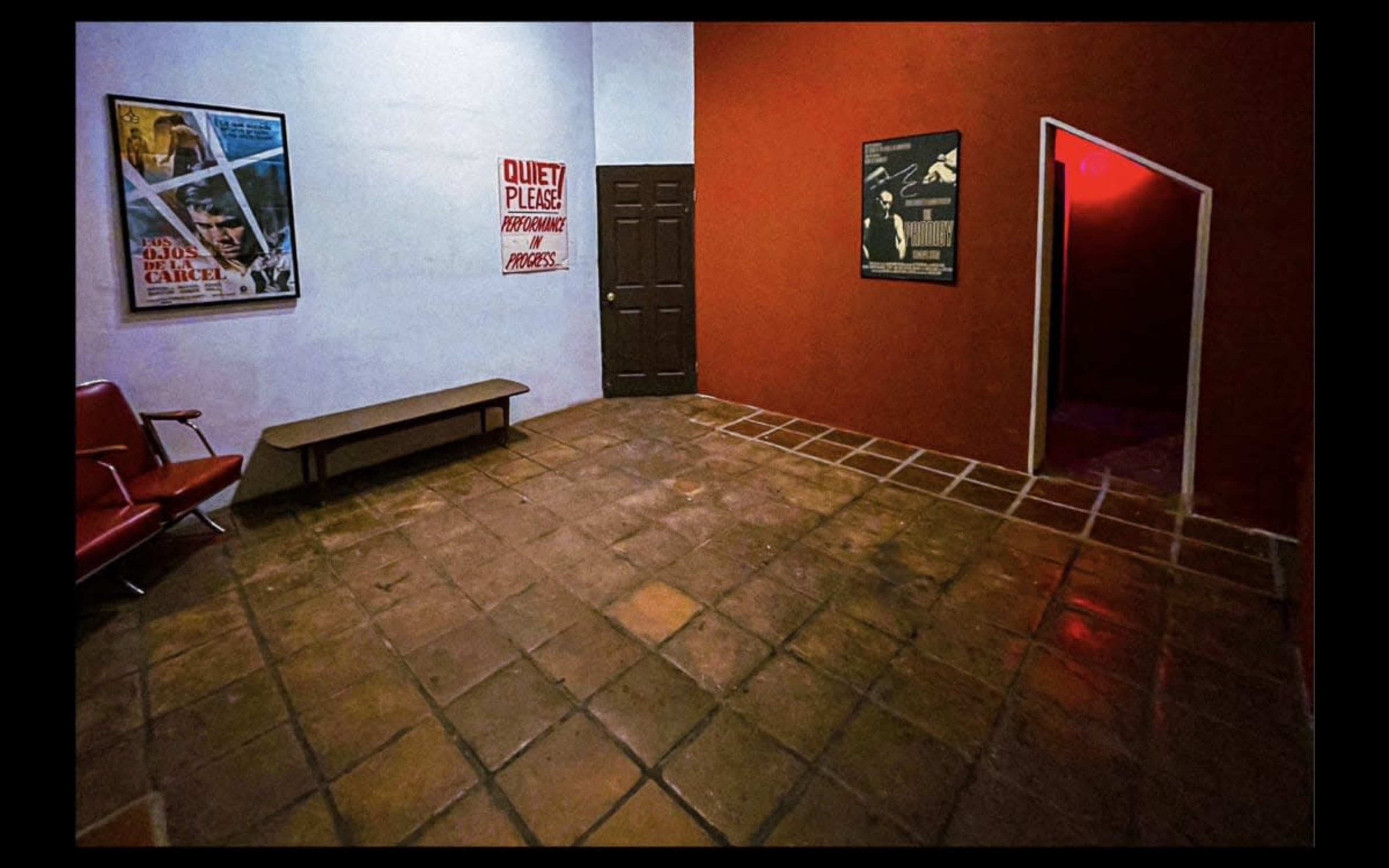 A dimly lit room with a red wall and a white wall, featuring a bench, posters on the walls, and a door leading to another area.