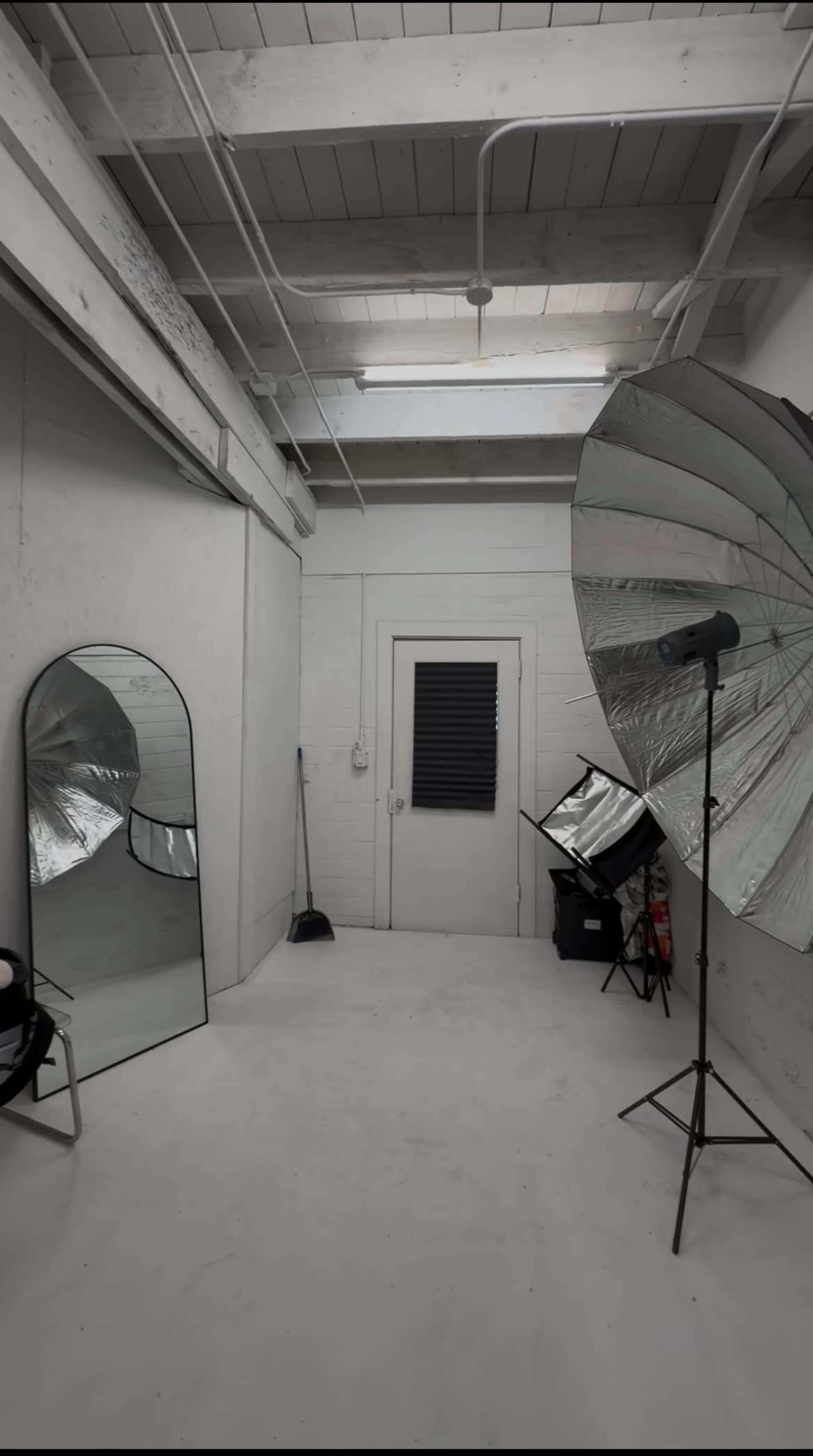 The image shows a photography studio with white walls, a large mirror, a light stand with an umbrella softbox, and a closed door at the back.