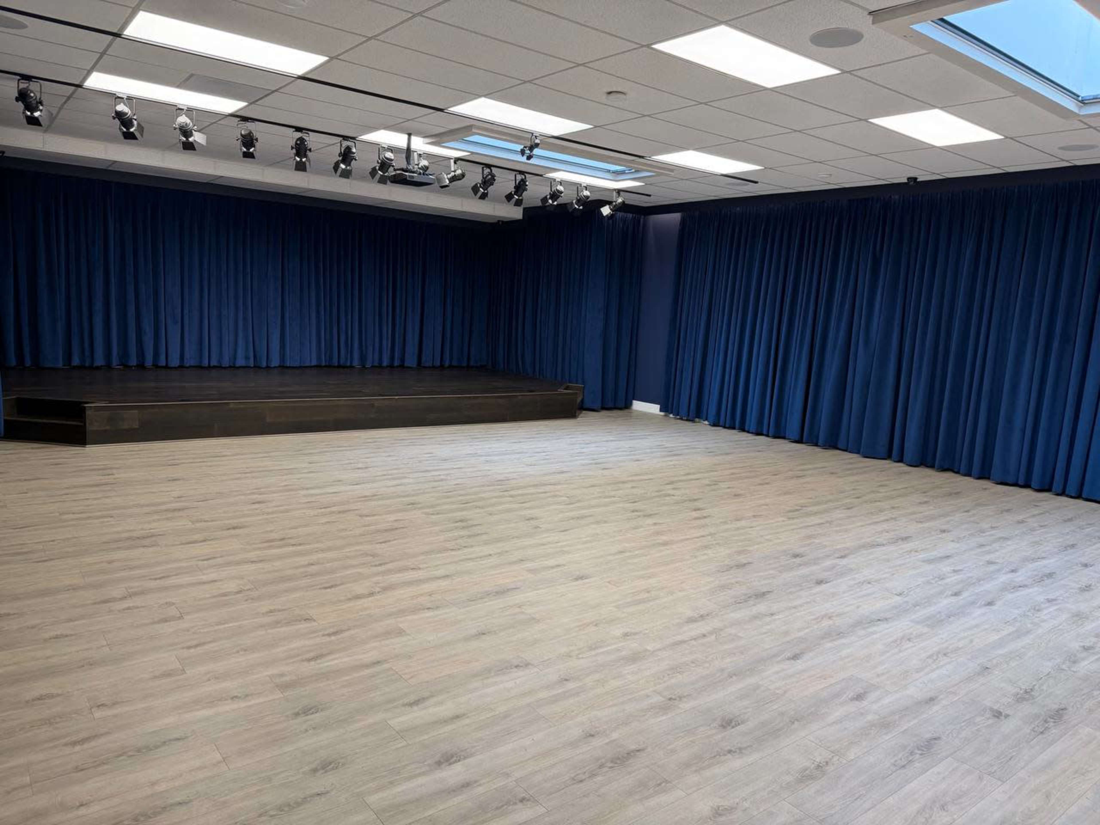 The image shows a spacious room with a wooden stage at the front, blue curtains lining the walls, and a light-colored wooden floor.