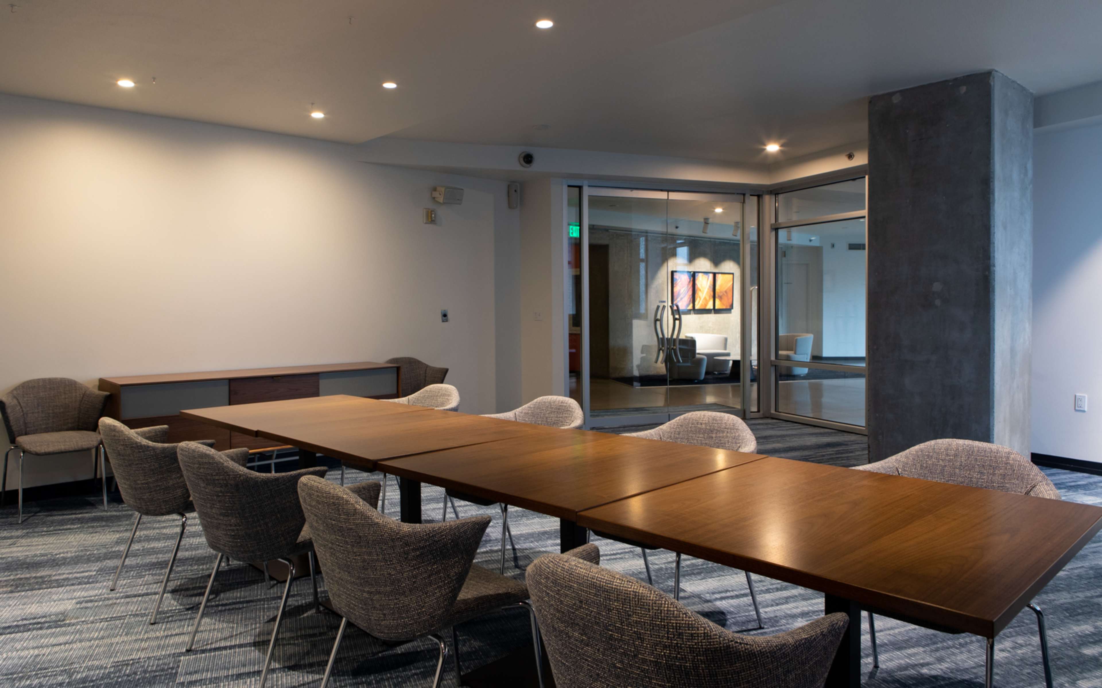 A long wooden table surrounded by eight chairs is set in a well-lit conference room, with a glass door leading to an adjoining area.