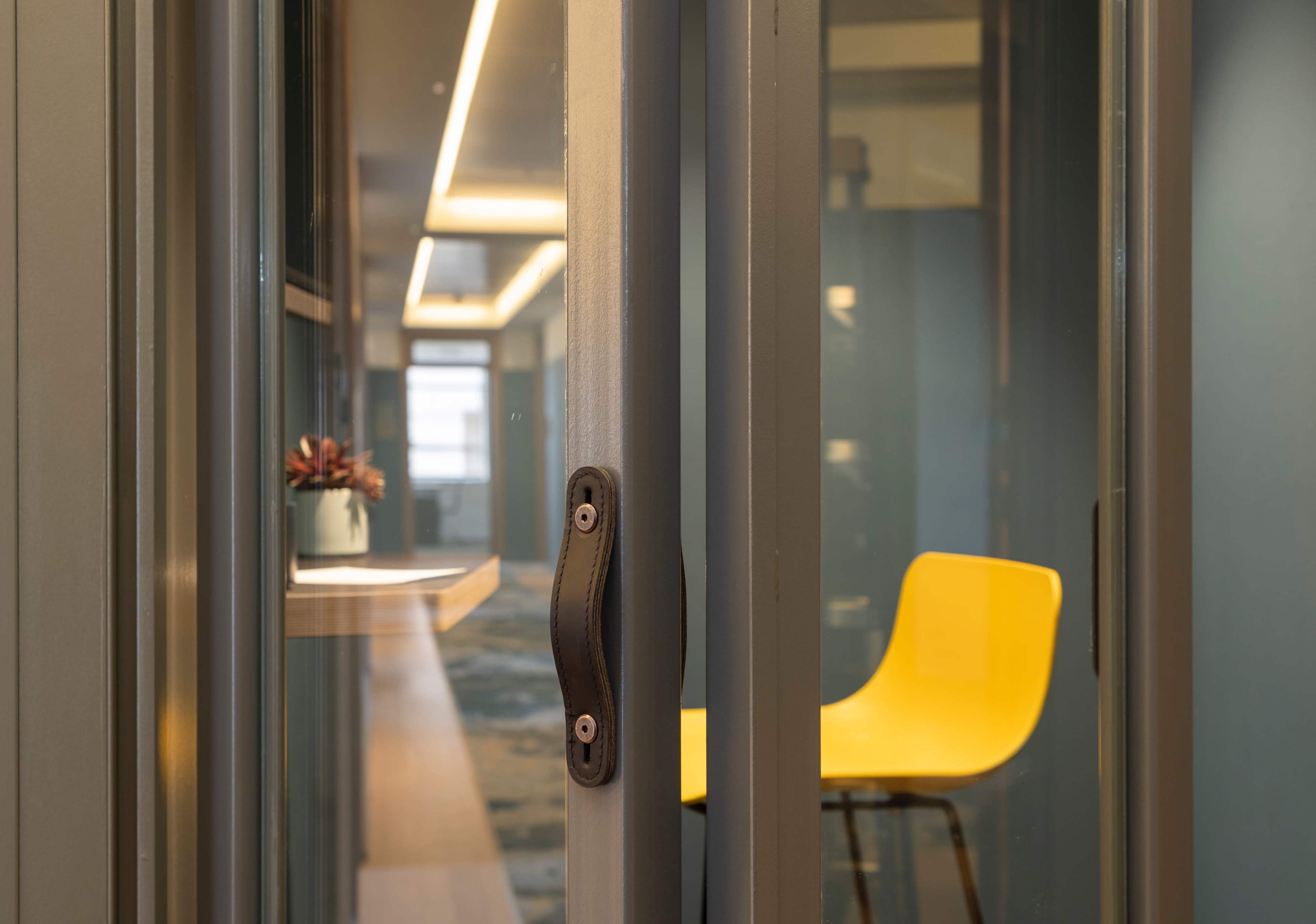 A yellow chair is positioned in a glass-walled meeting room with soft lighting and a corridor visible in the background.