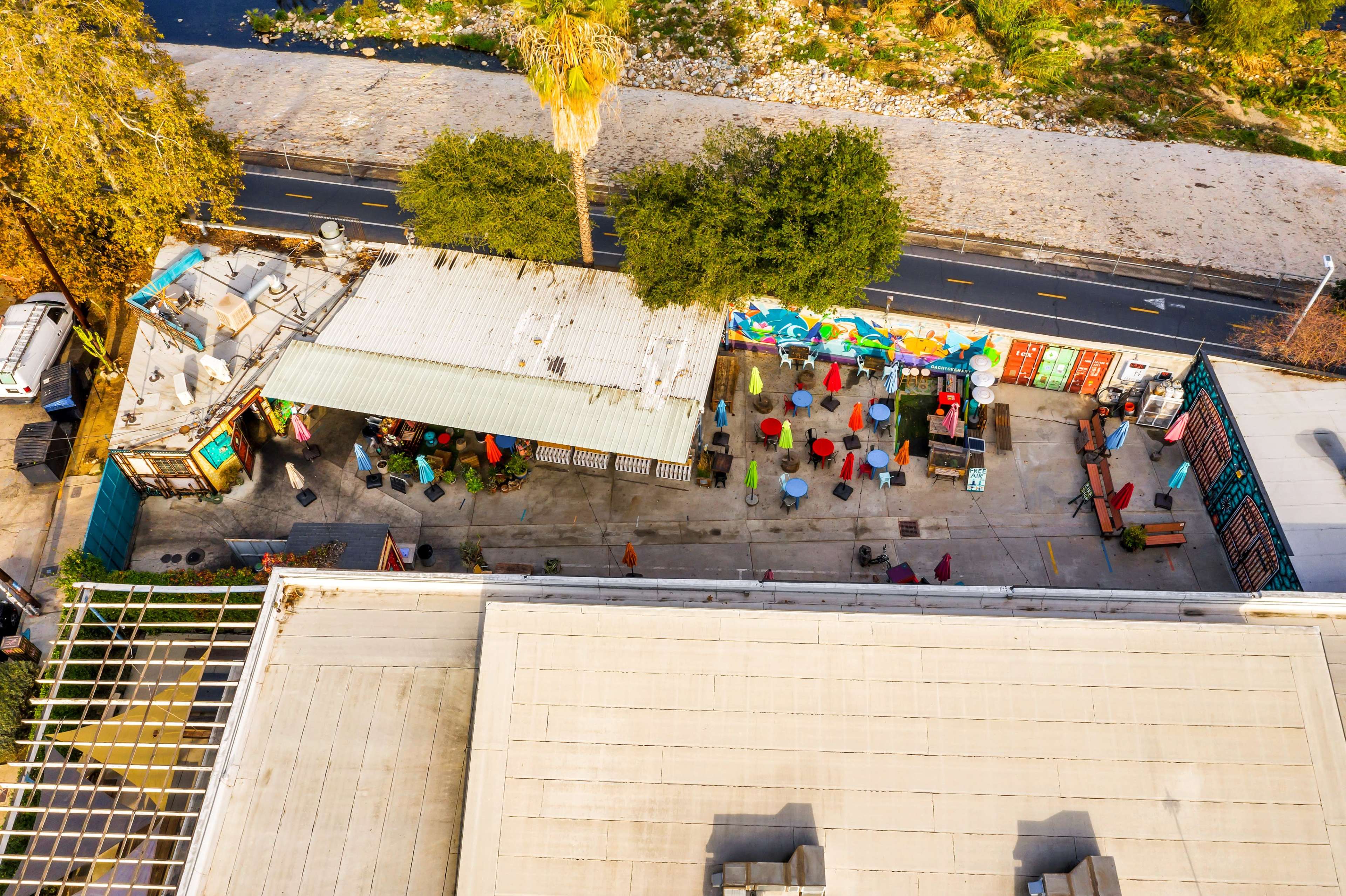An aerial view of a colorful outdoor dining area with umbrellas, adjacent to a riverbank and a road.