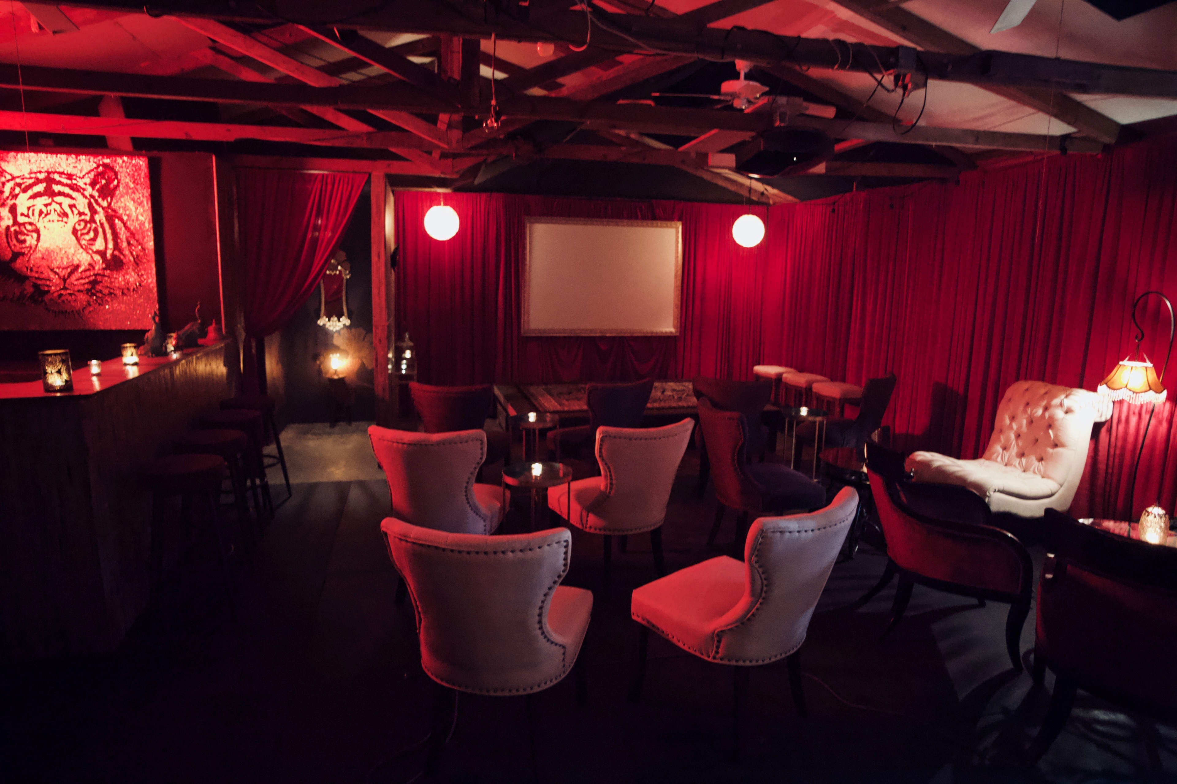 A dimly lit lounge area with red curtains, various chairs, a bar counter, and soft lighting.