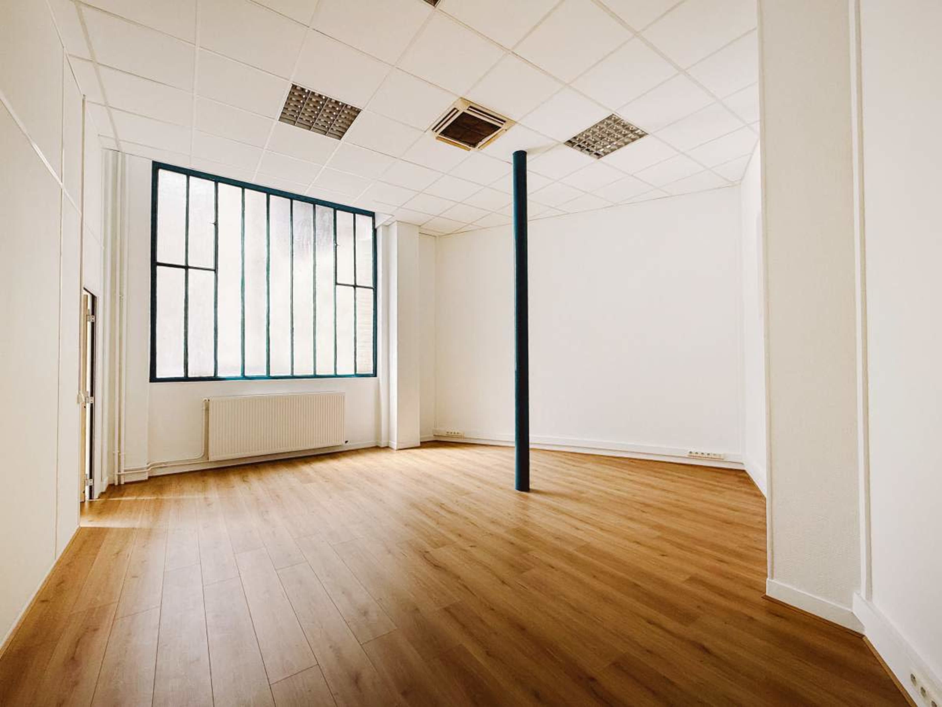 An empty room with wooden flooring, a tall window on one wall, and a vertical pillar in the center.