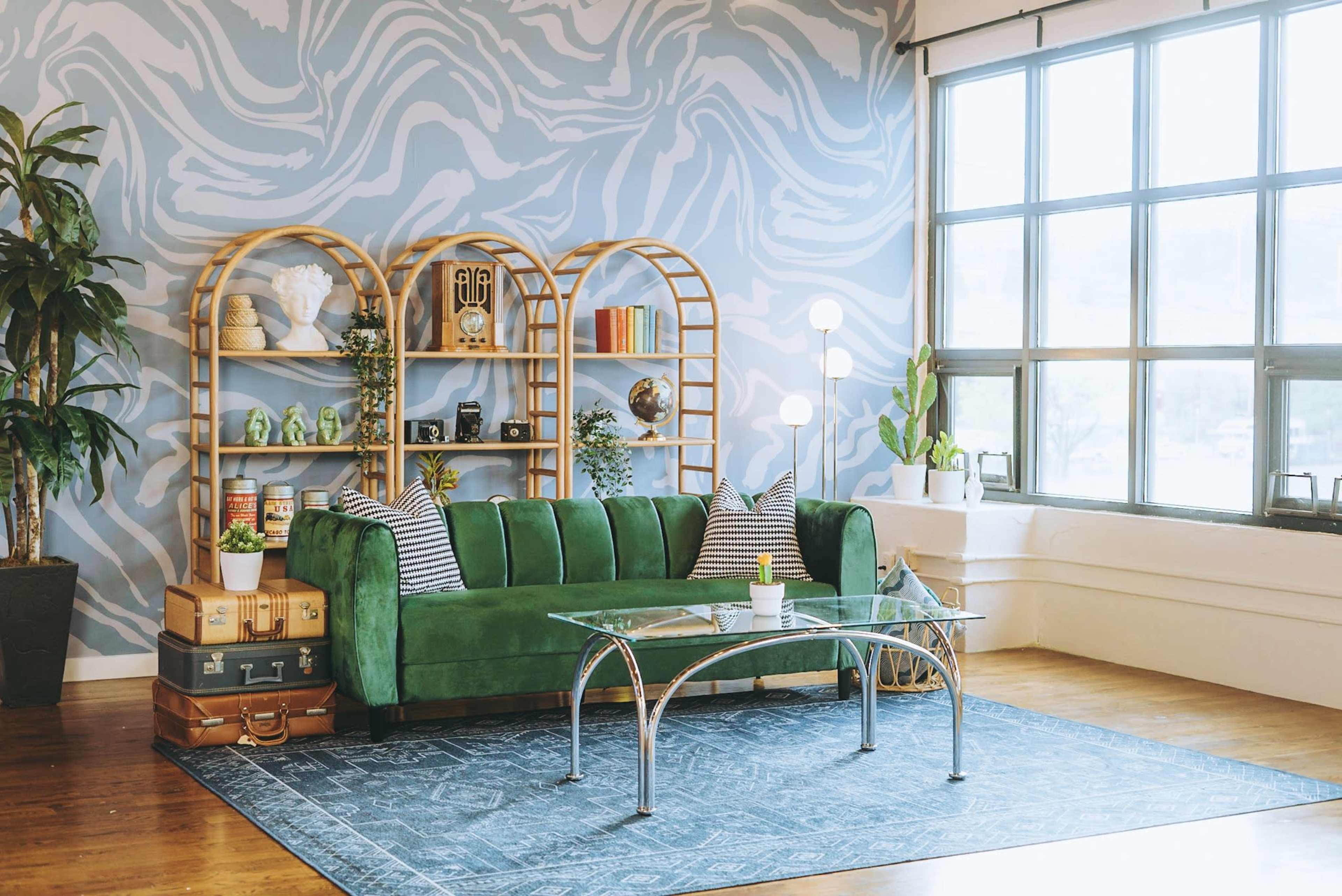 A green velvet sofa is placed in front of a large window with a blue abstract wall design, accompanied by a glass coffee table and a bookshelf filled with decorative items and plants.
