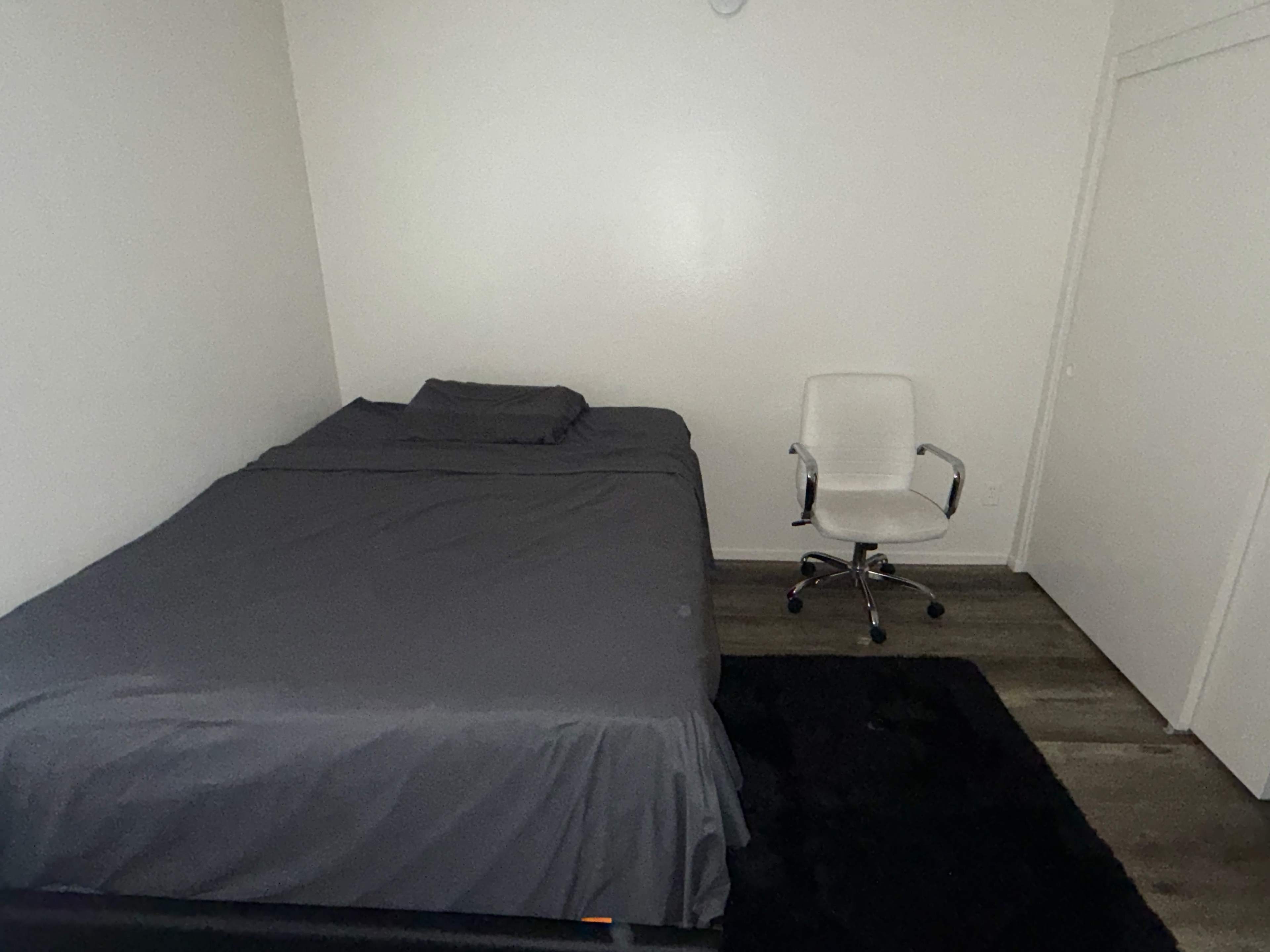 The image shows a simple bedroom with a bed covered in gray bedding and a white office chair beside it.