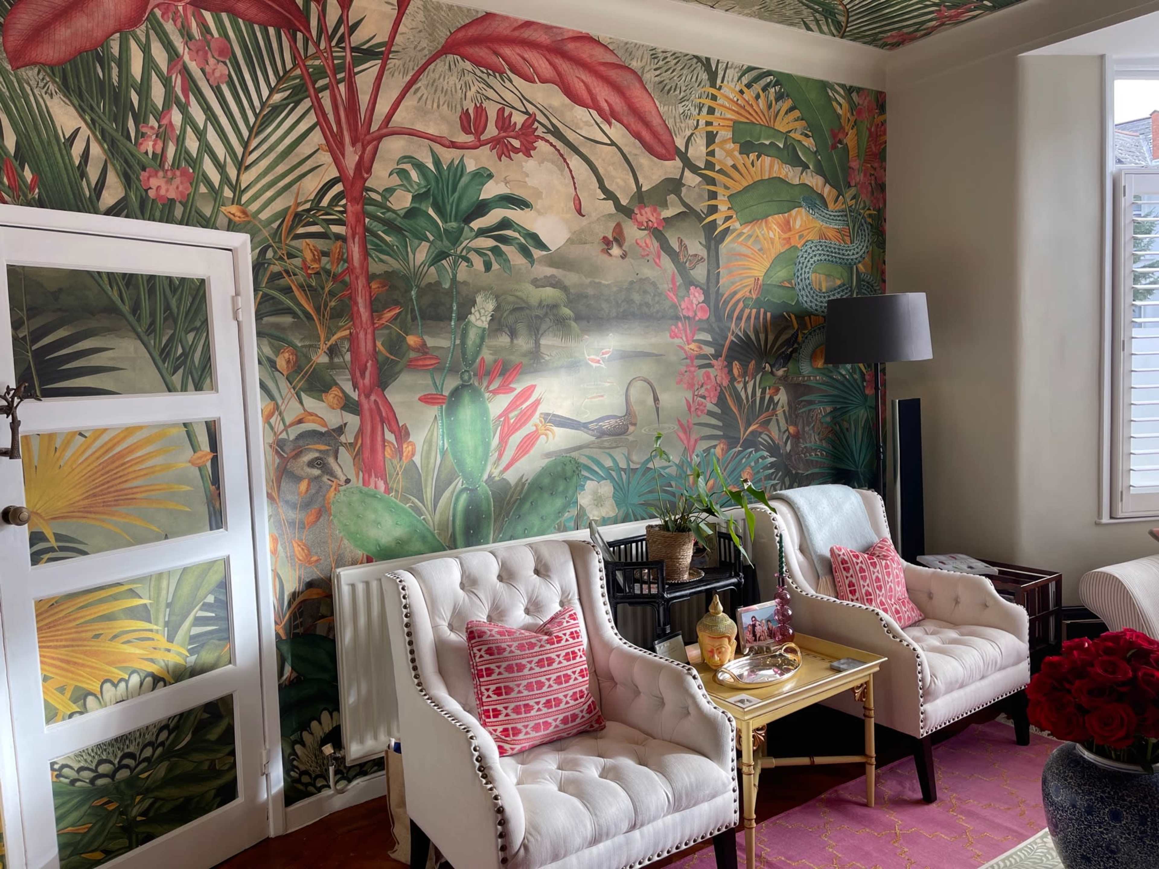 The image shows a living room with two upholstered chairs facing a tropical mural featuring various plants and a landscape.