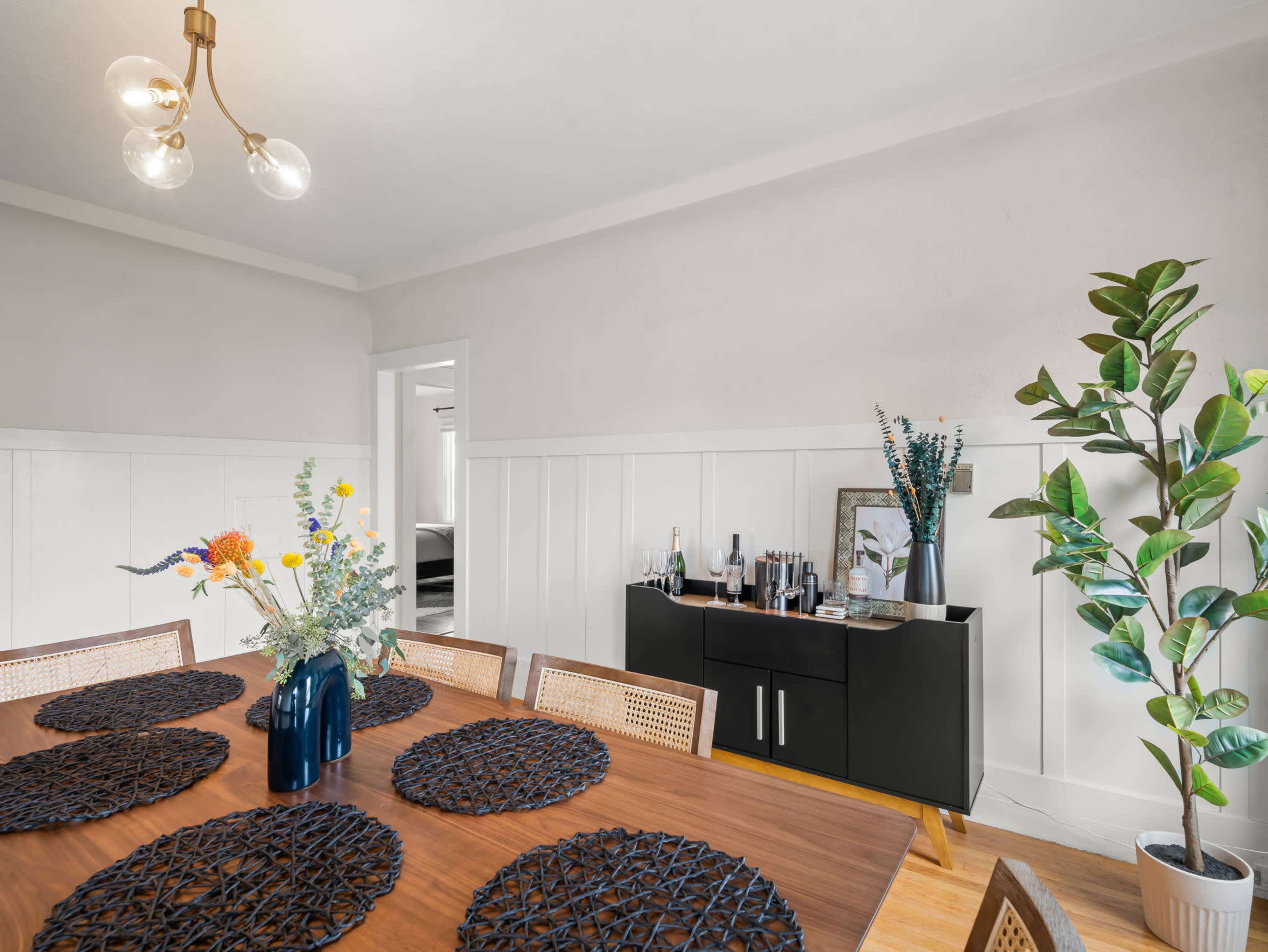 A dining area features a wooden table with dark round placemats, a vase of flowers, and a sideboard displaying various decorative items and a mirror.